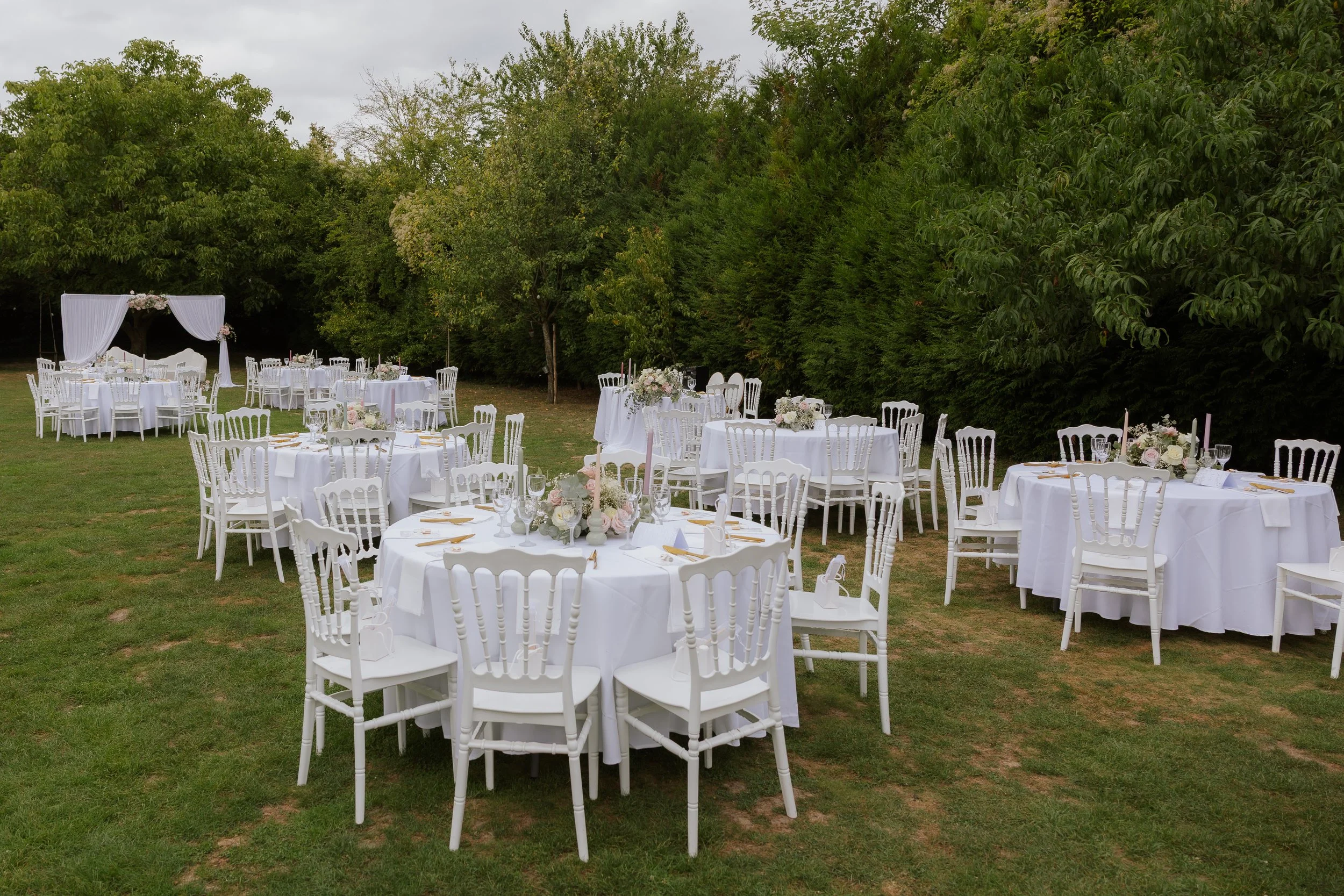 Tables rondes avec nappes blanches, vaisselle et arrangements floraux dans un jardin pour un événement ou mariage, avec un dais blanc décoré de fleurs en arrière-plan.