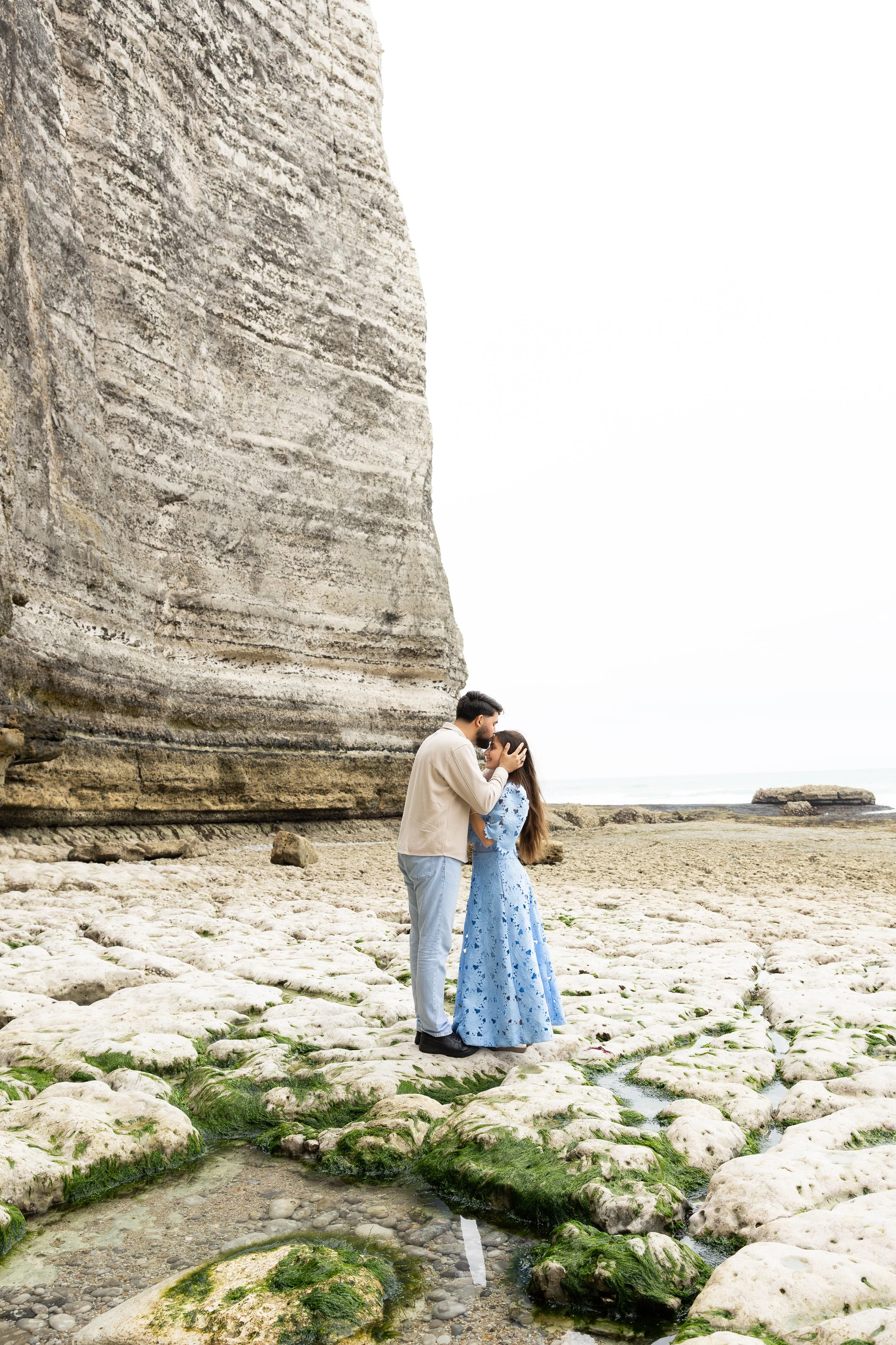 Un couple se tenant par la main et se regardant tendrement près de la mer, avec une grande falaise rocheuse en arrière-plan, en journée brumeuse.