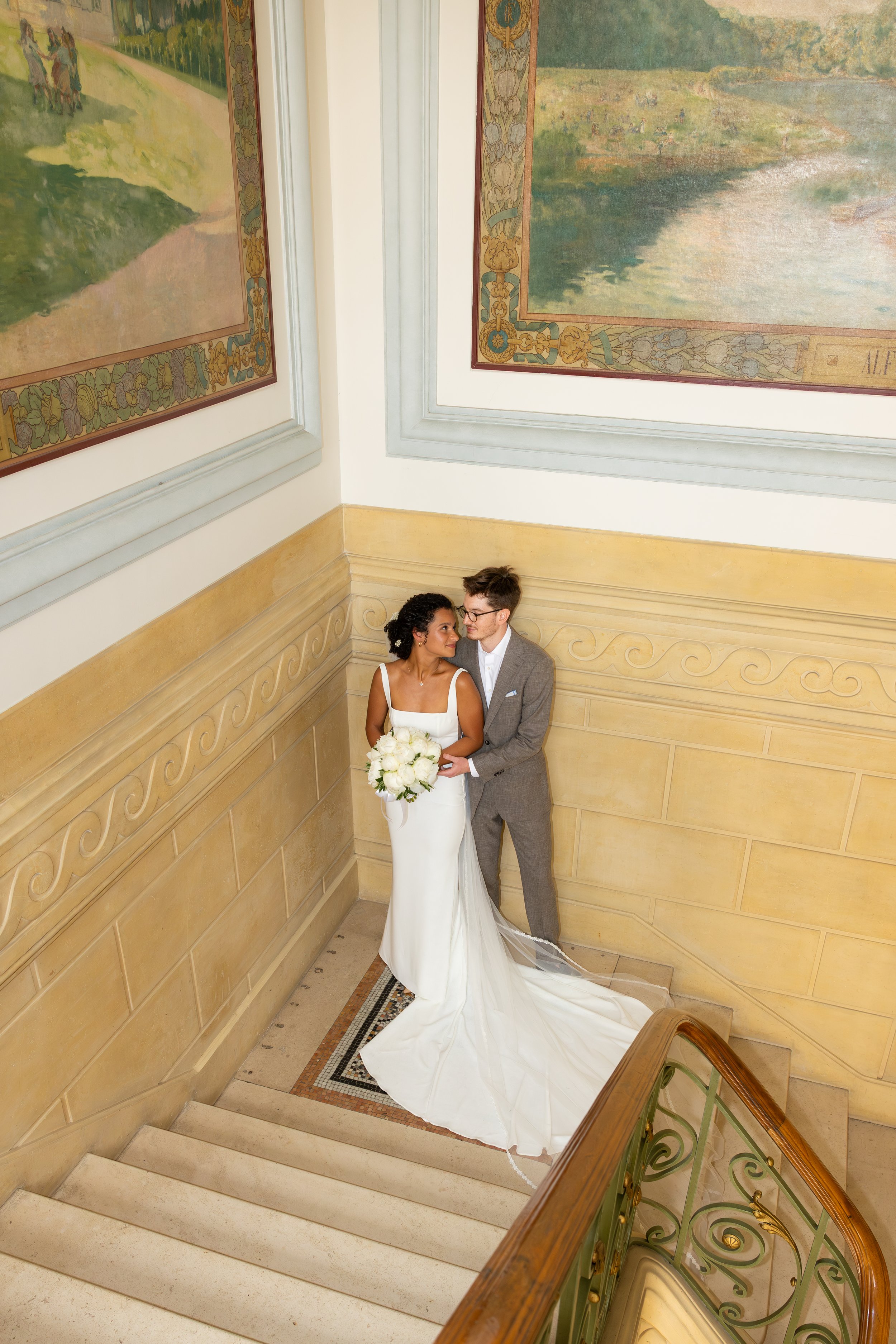 Un couple de mariés debout dans un escalier, à la mairie de Maison Alfort, la femme en robe blanche avec un bouquet de fleurs blanches, l'homme en costume gris, dans un décor élégant avec des peintures murales.