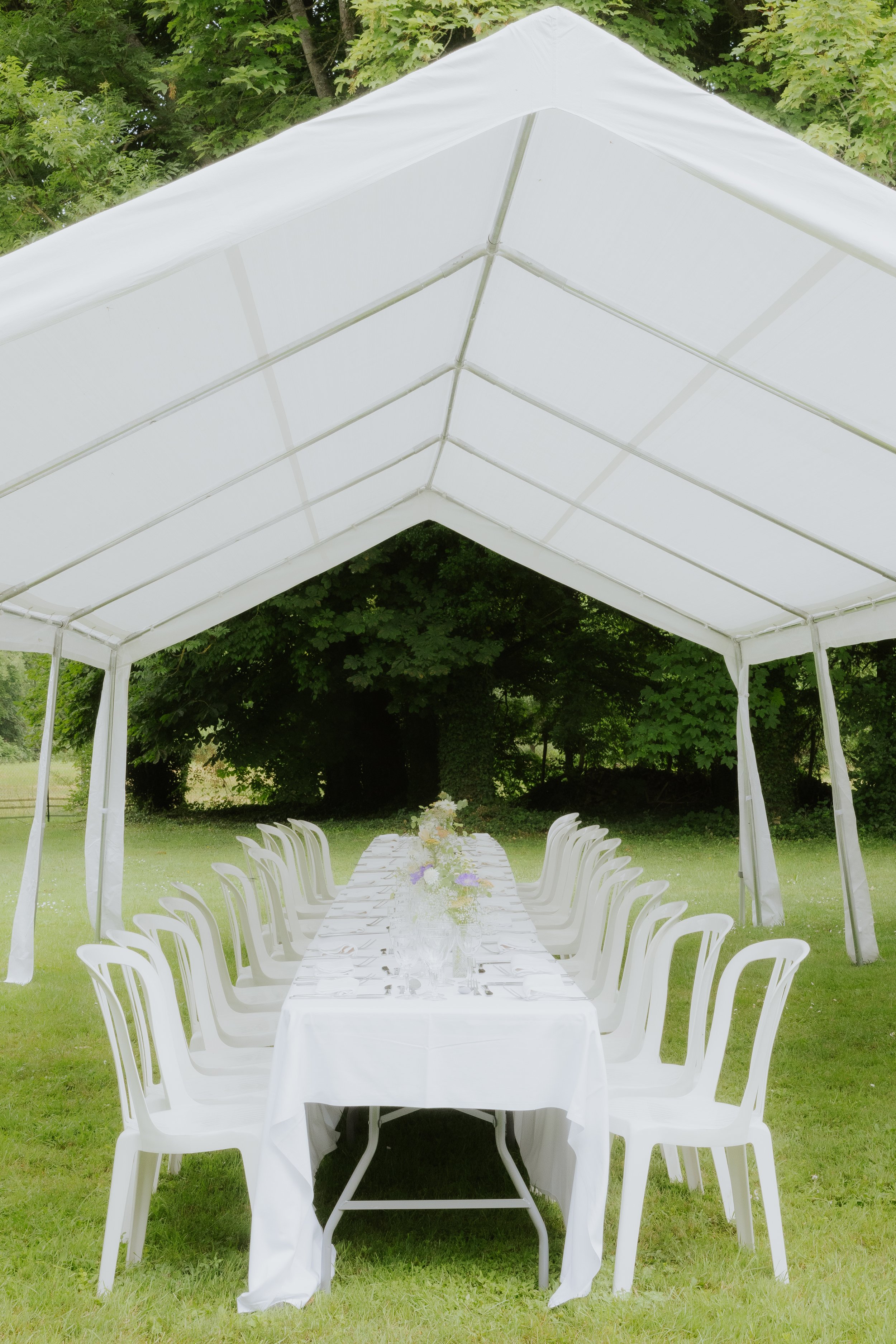 Une table de banquet en plein air sous une tente blanche, entourée de chaises en plastique blanc, avec des fleurs en centre de table et un fond de verdure dense.