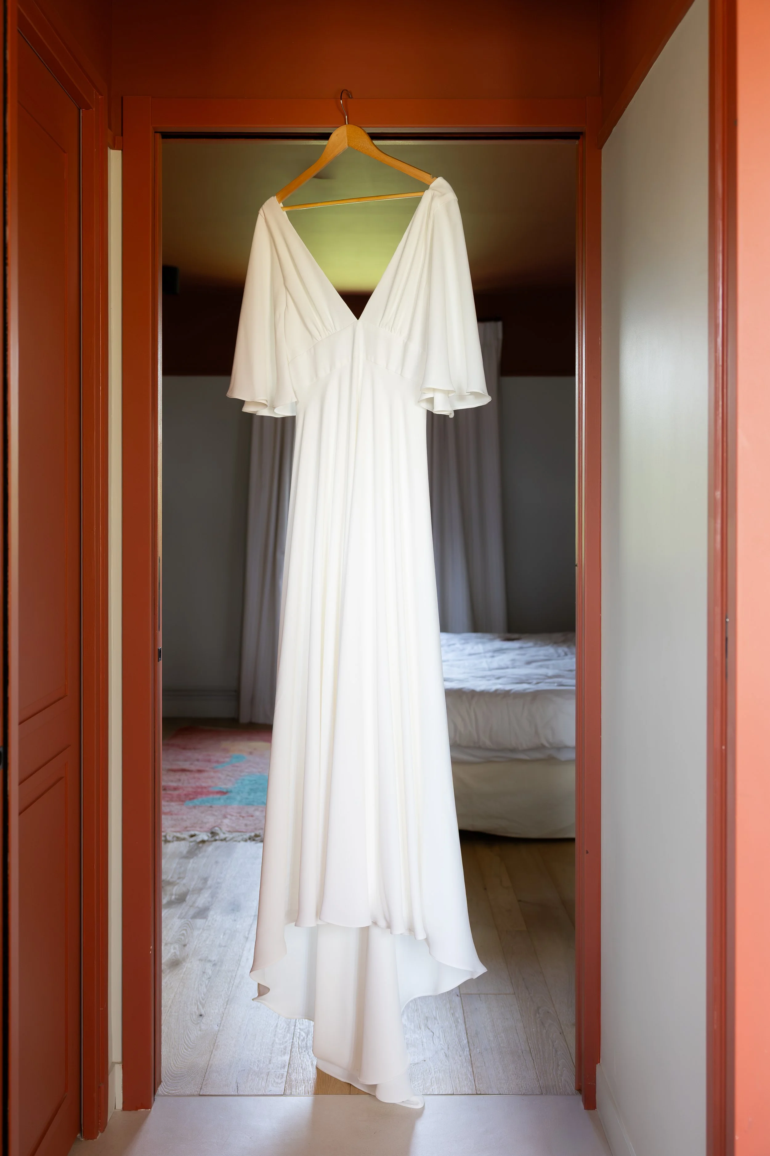 Robe de mariage blanche suspendue sur un cintre dans une entrée, sur une porte en bois rouge, avec une chambre en arrière-plan.