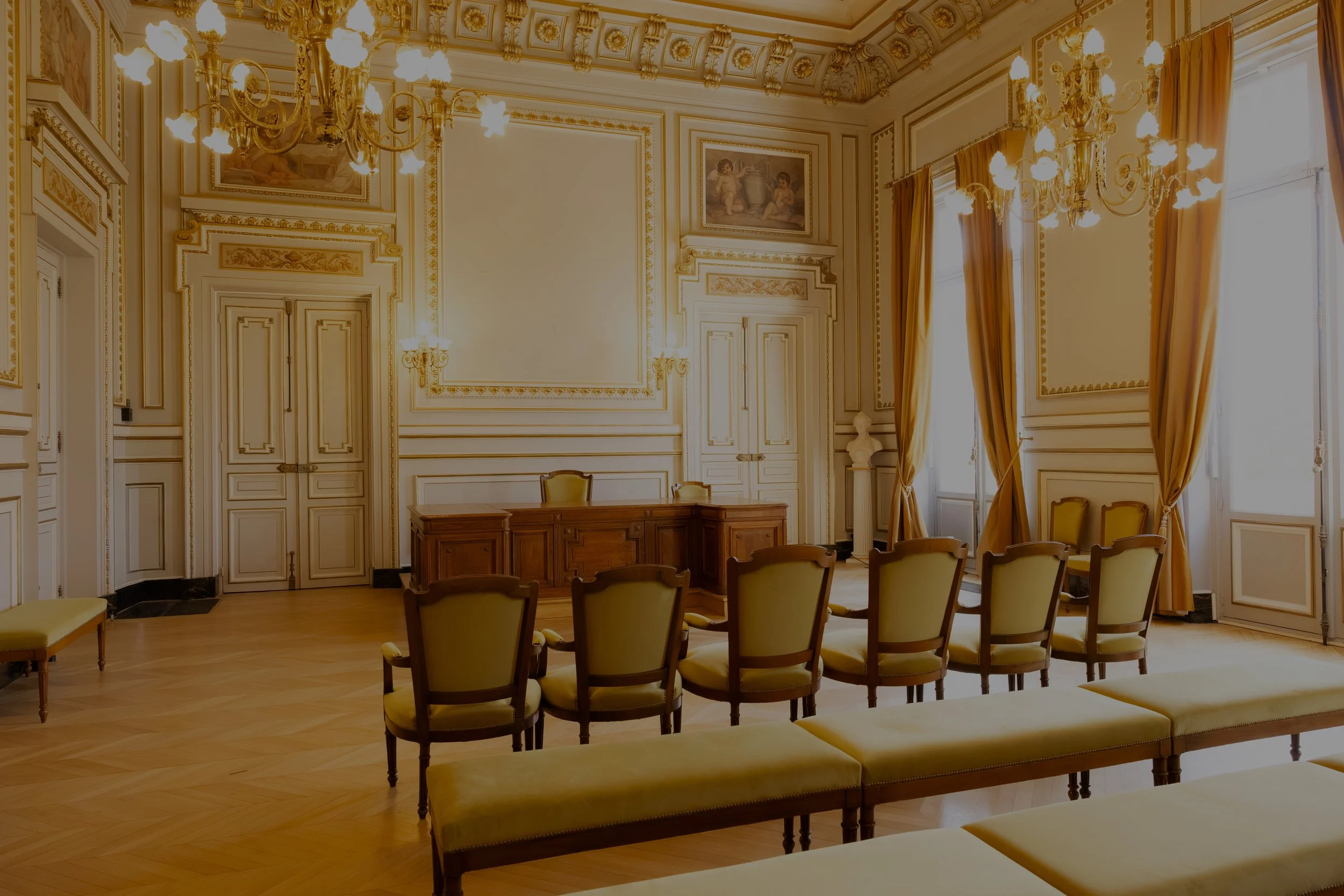 Salle de mariage Mairie de Clichy, luxueuse avec des chaises en bois et tissu beige, un bureau en bois, des grandes fenêtres avec des rideaux dorés, un lustre en cristal, des murs ornés de moulures et de peintures anciennes.