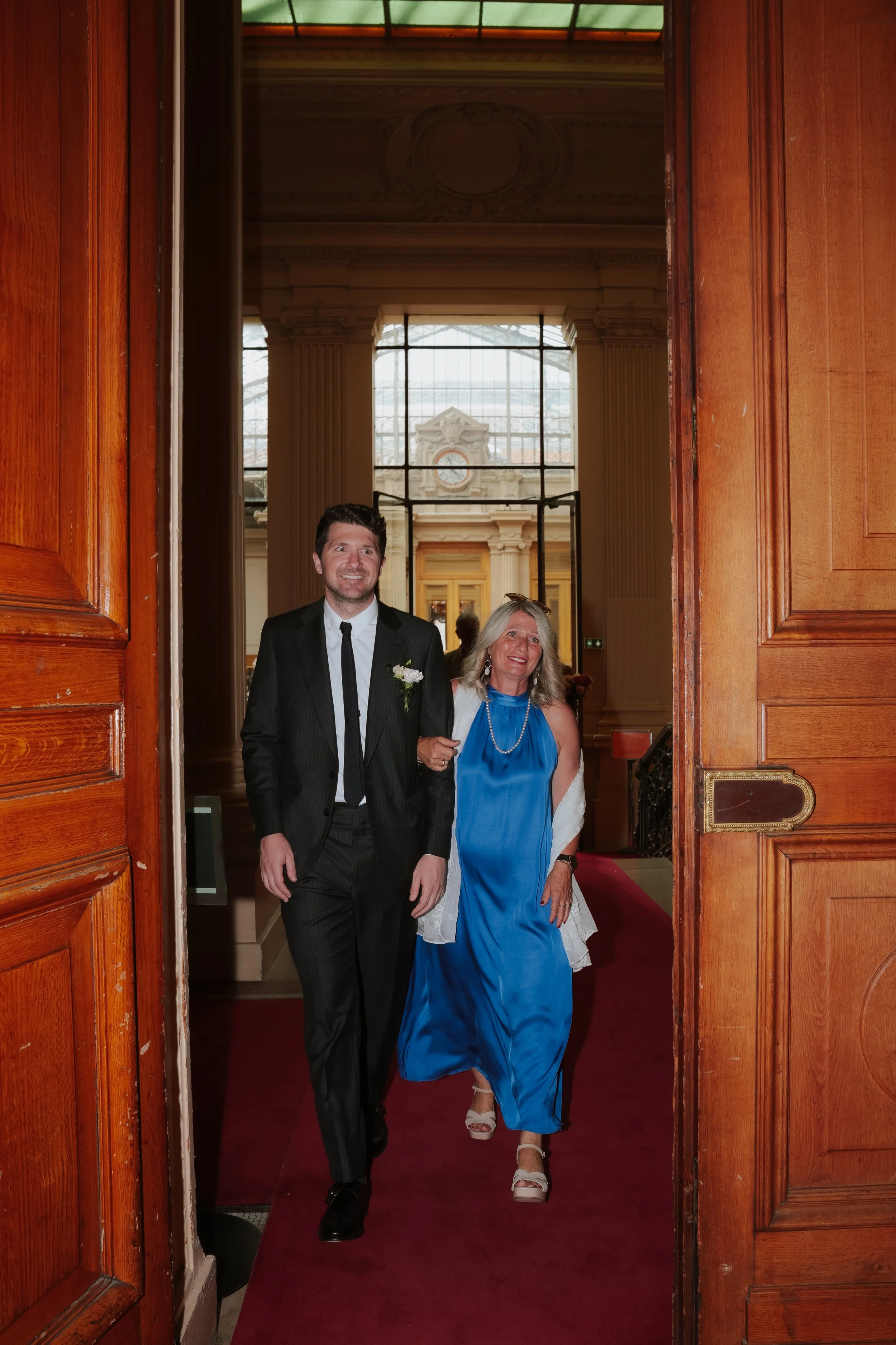 Un homme en costume noir et une femme en robe bleue marchent côte à côte dans un bâtiment élégant avec des colonnes et une horloge sur le mur en arrière-plan.