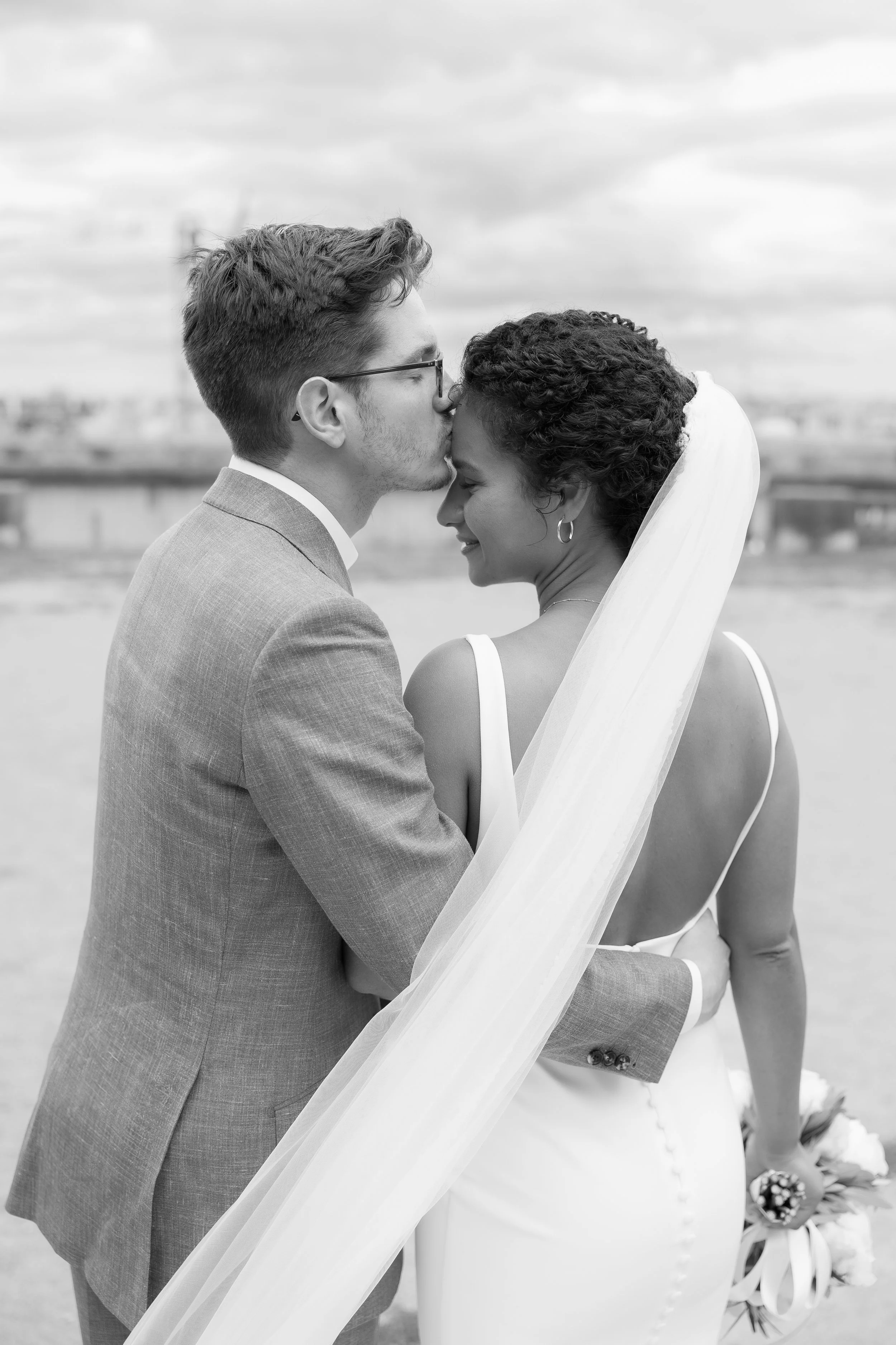 Un couple de mariés en costume élégant, l'homme embrassant la mariée sur le front, tenant un bouquet, en extérieur, en noir et blanc.