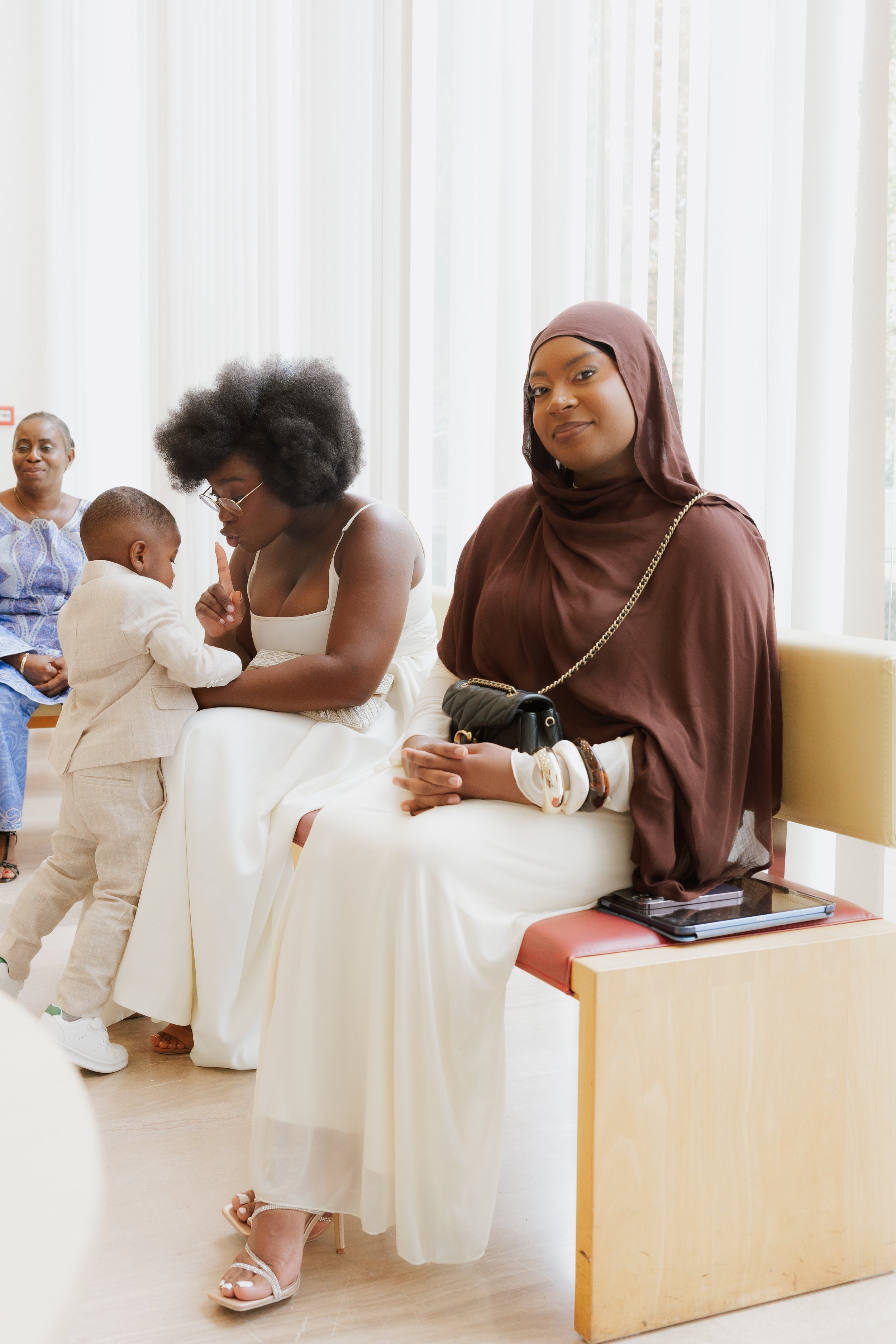 Deux femmes assises dans une salle, une d'elles souriant et portant un hijab marron, l'autre parlant à un jeune garçon. Deux autres femmes sont visibles en arrière-plan.