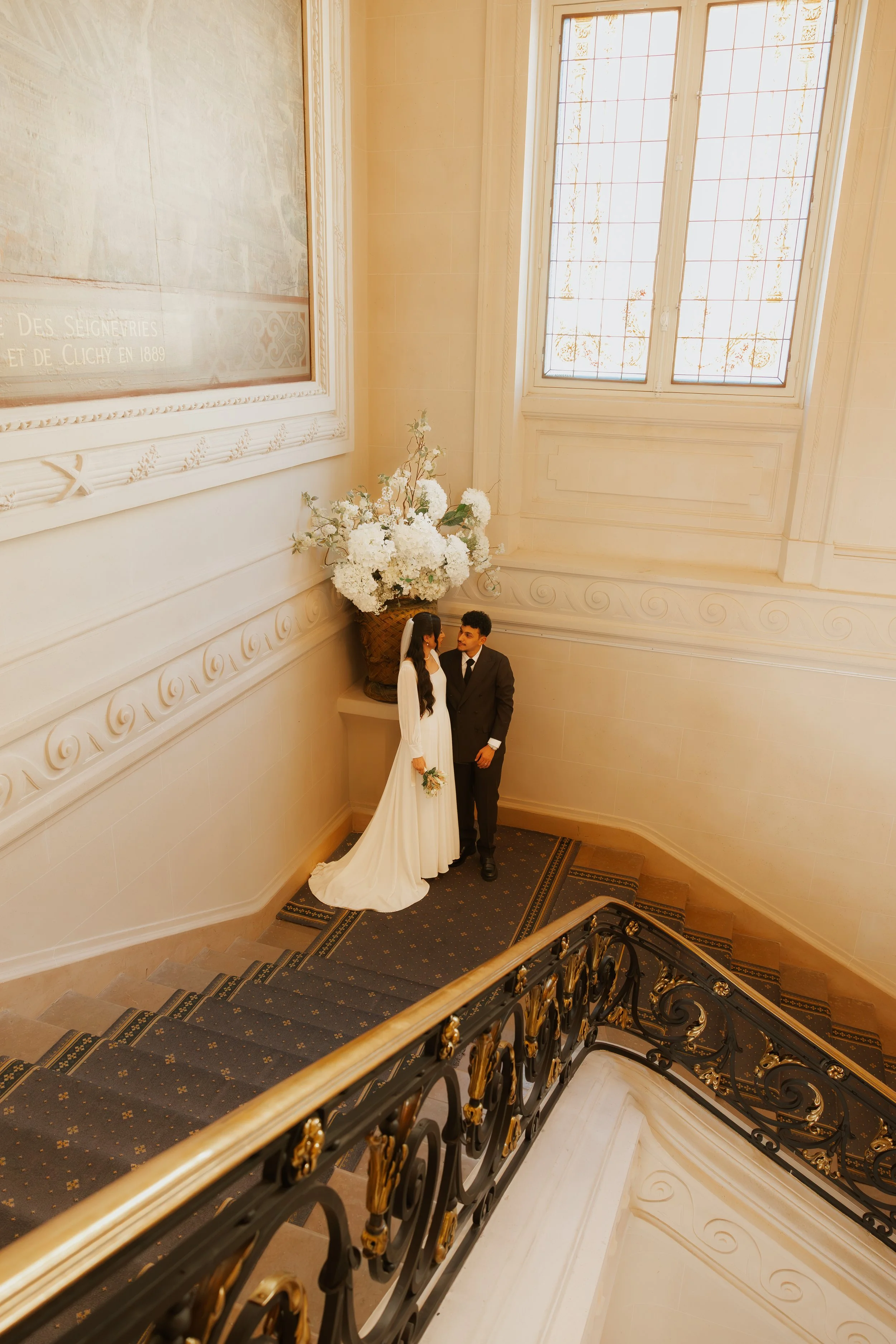 Un couple en vêtements de mariage, la femme en robe blanche et le mari en costume noir, se tiennent dans un couloir élégant avec une grande fenêtre et un grand vase de fleurs blanches sur un rebord.