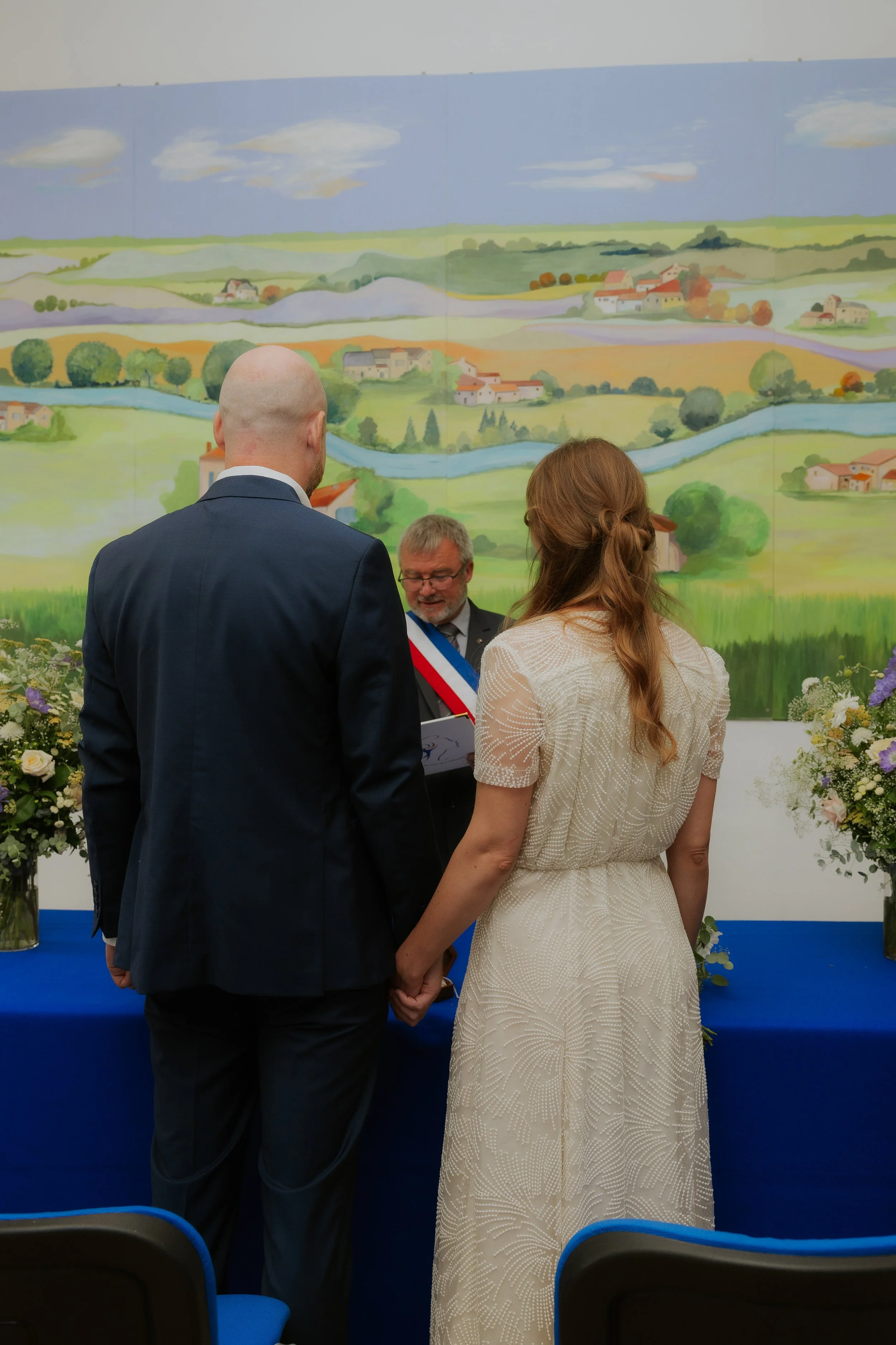 Un couple tenant la main lors d'une cérémonie officielle, face à un officiel avec la collation tricolore, avec un tableau représentant un paysage rural en arrière-plan.