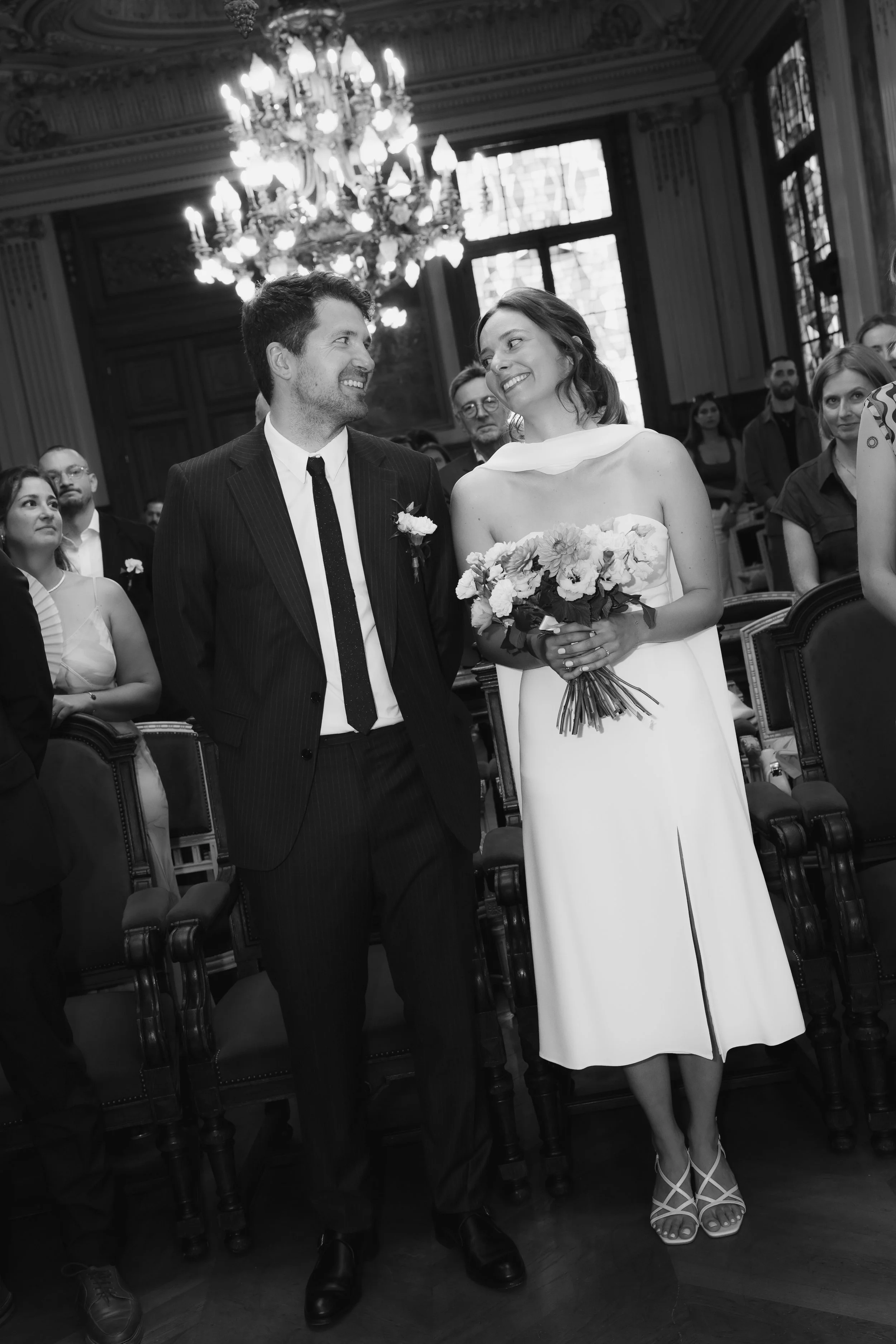 Un couple lors d'une cérémonie de mariage dans une salle élégante, la mariée en robe blanche tenant un bouquet de fleurs, le marié en costume noir, entourés d'invités assis et debout.