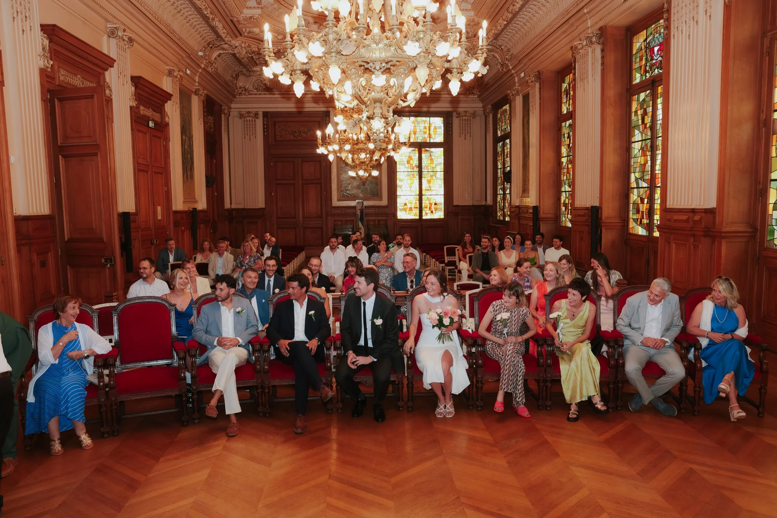 Une célébration de mariage, à la mairie du 18ème arrondissement de Paris dans une salle élégante avec un plafond en bois, vitraux colorés, chandelier imposant. Le couple de mariés est assis au centre, entouré de leurs invités souriants.