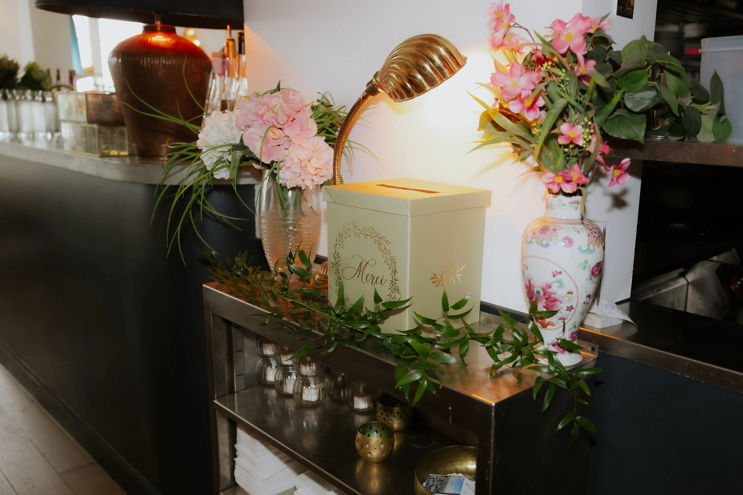 Vases avec des fleurs roses et blanches, une boîte à offrir blanche avec le mot 'Merci', un éclairage doux, un lampadaire en métal, et un environnement décoratif élégant.