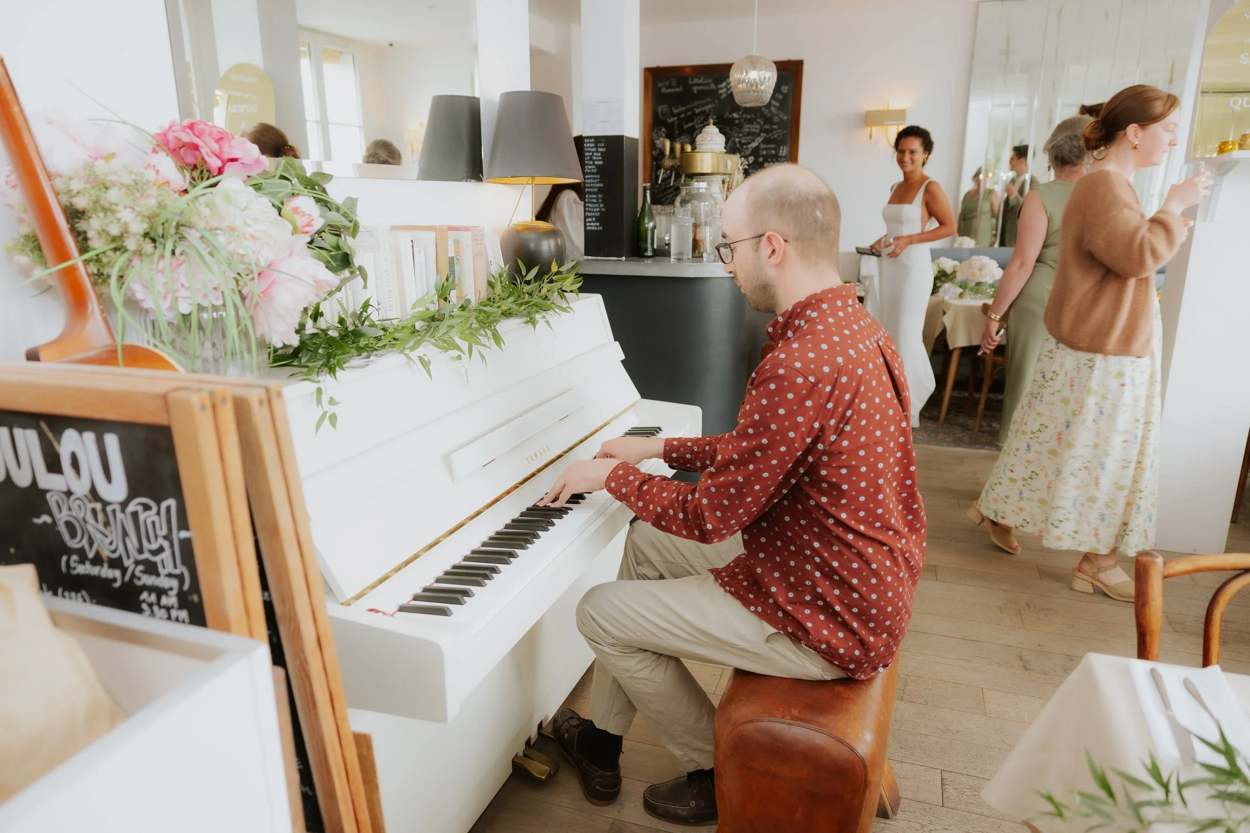 Un homme joue du piano blanc dans un café ou un restaurant, décoré avec des fleurs et des livres, avec plusieurs personnes en arrière-plan