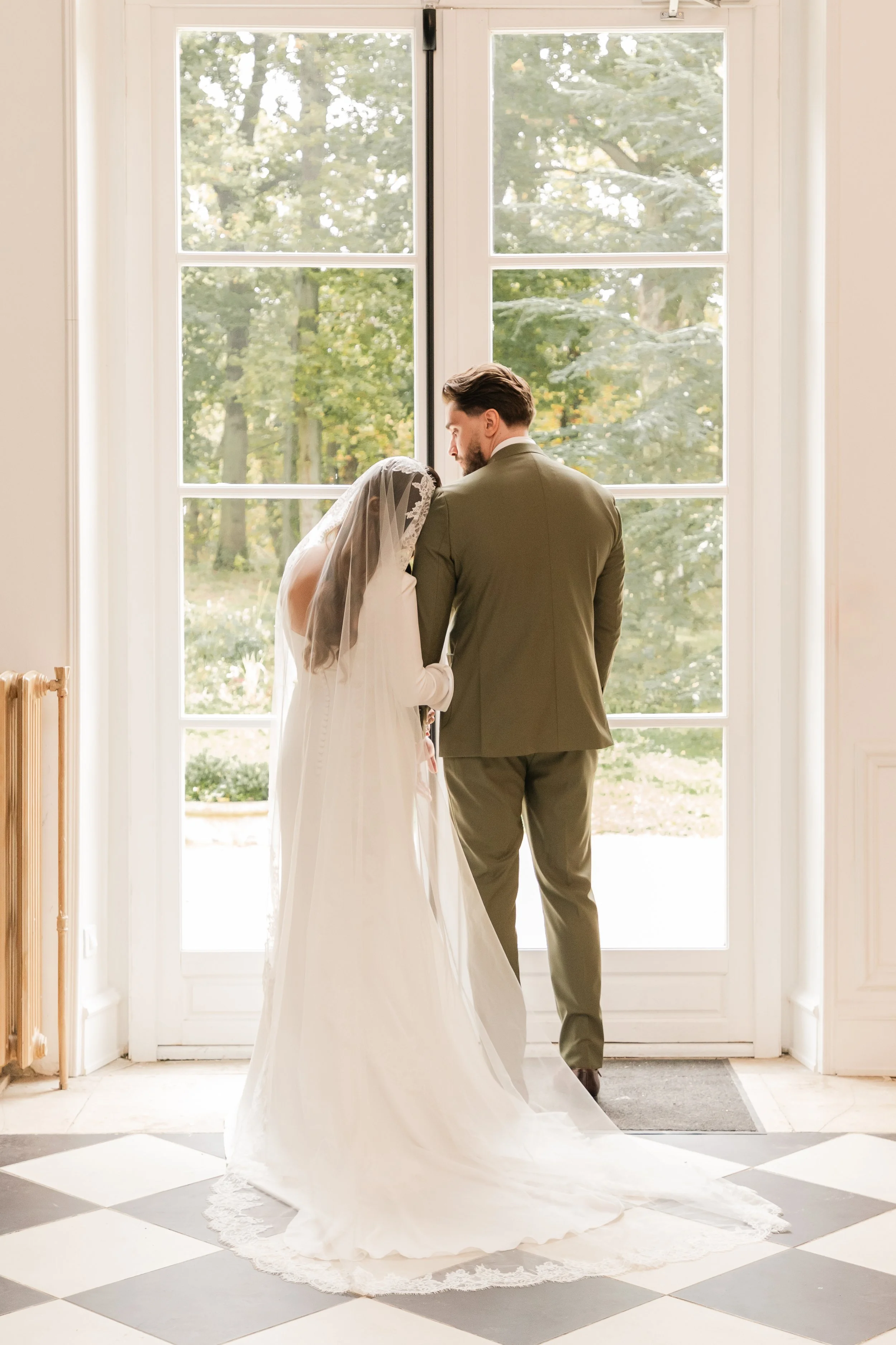 Un couple de mariés debout devant une grande fenêtre, la mariée en robe blanche avec une voile et le marié en costume vert, à l'intérieur d'une pièce lumineuse.