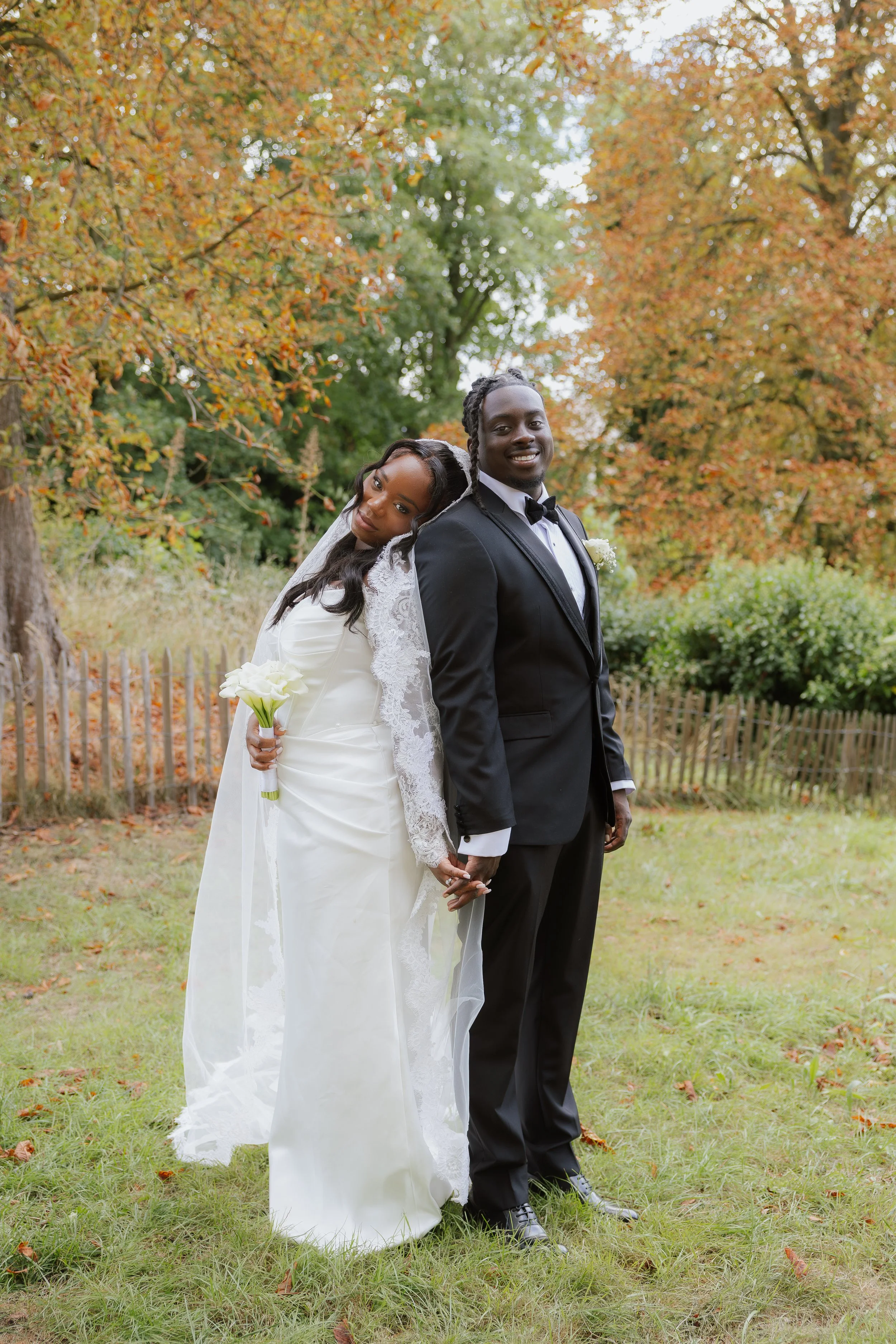 Un couple de mariés pose dans un parc aux arbres aux feuilles d'automne. La mariée porte une robe blanche et un voile, tenant un bouquet blanc, et le marié porte un costume noir avec un nœud papillon.