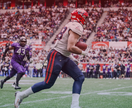 An American football player in a red and gray uniform running with the football during a game, with a crowd in the background.