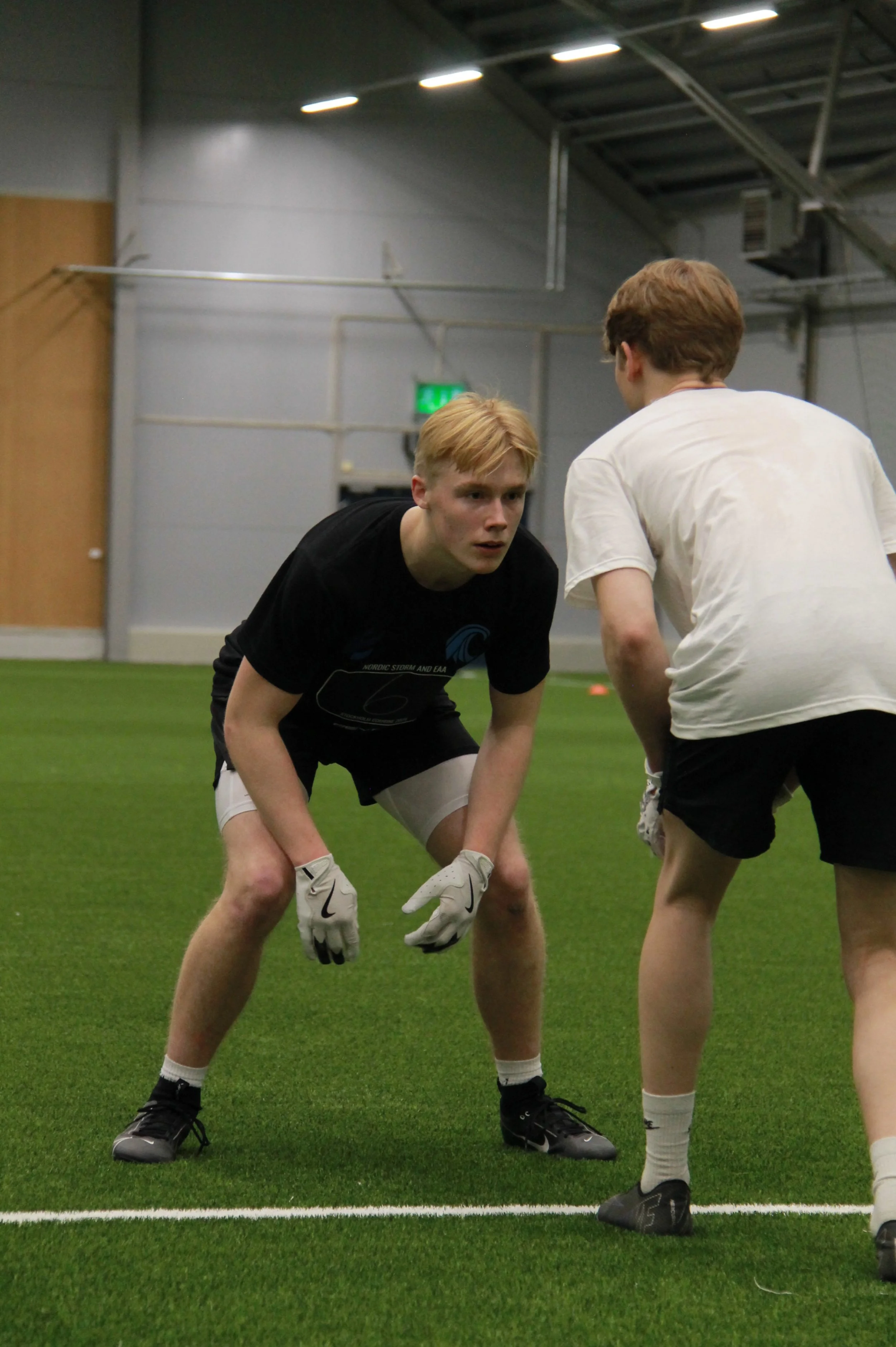 Two young men practicing football drills indoors on a green turf field.