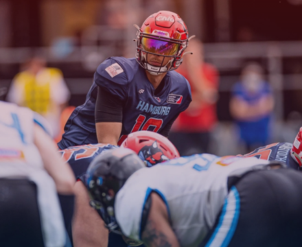 An American football player in a navy blue uniform and red helmet with a face shield, preparing to hike the ball during a game.