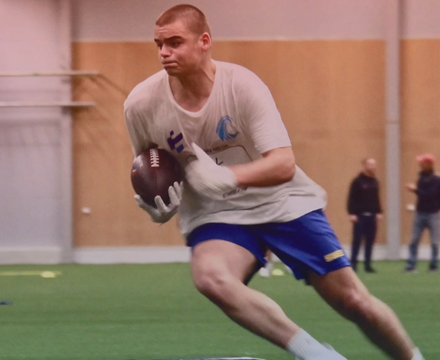 A young man running indoors while holding a football, wearing a gray t-shirt and blue shorts.