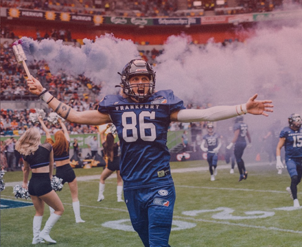 An American football player wearing a blue jersey with the number 86, celebrating on the field with arms outstretched, surrounded by cheerleaders and fans, smoke in the background at a stadium.
