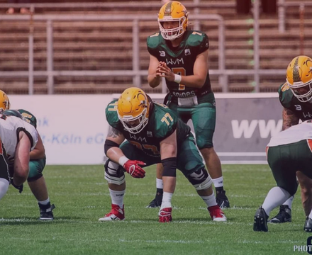 American football players in green and yellow uniforms are lined up on the field during a game, with the quarterback holding the ball.