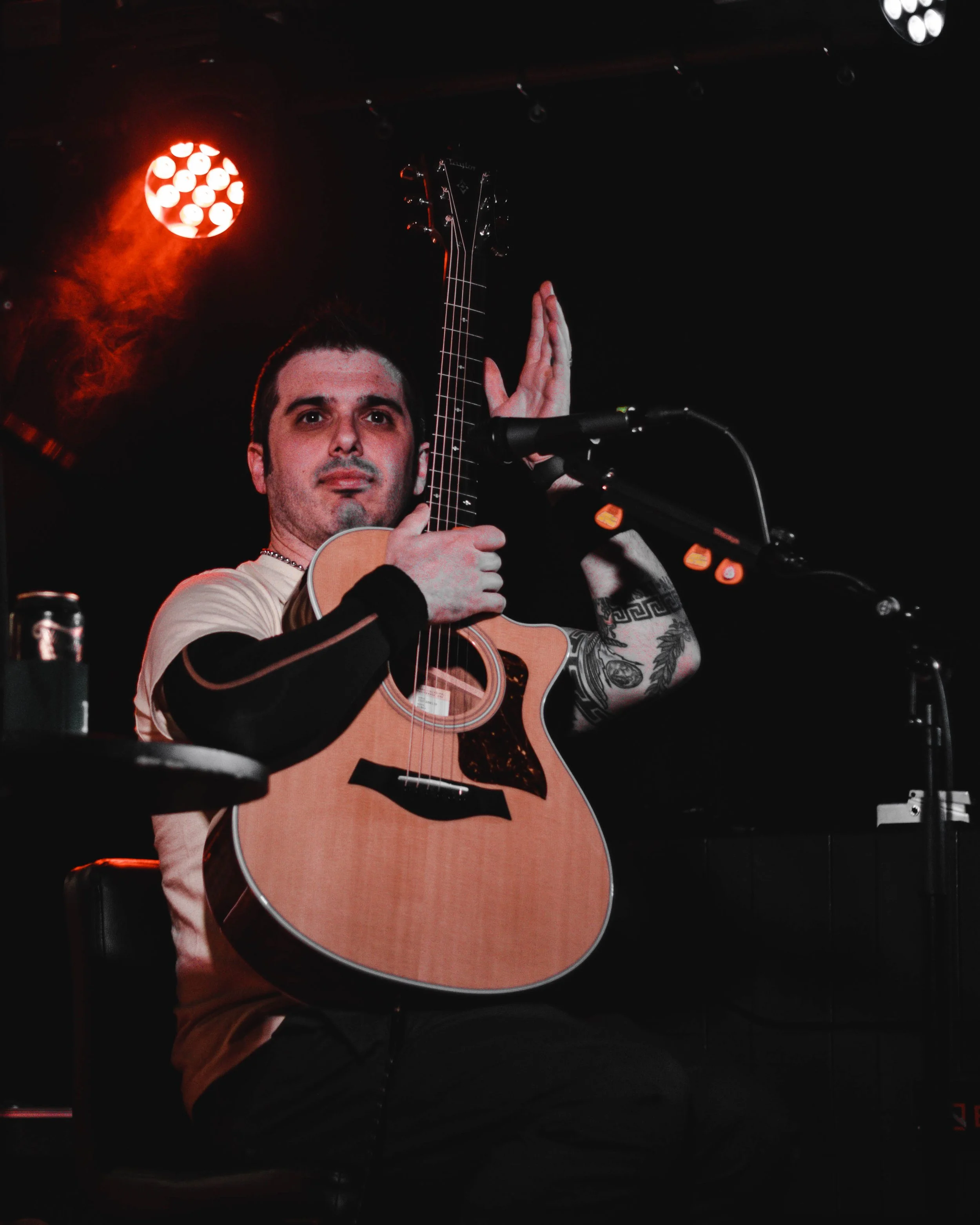 A man with short dark hair and tattoos on his arm sitting on a chair on stage, holding an acoustic guitar, with stage lights overhead.