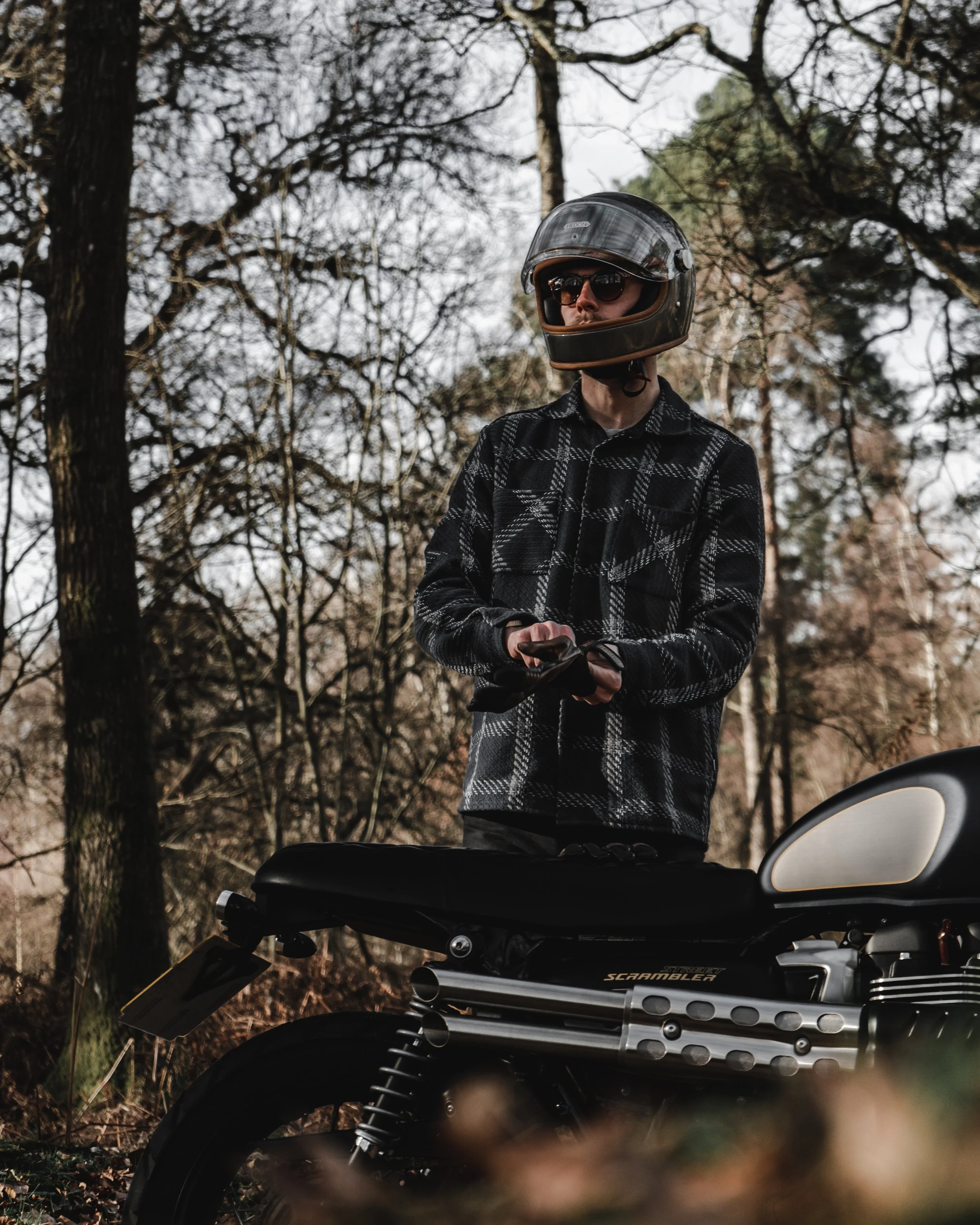 Person standing outdoors in a wooded area wearing a black helmet and sunglasses, dressed in a black and white plaid jacket, next to a black motorcycle.