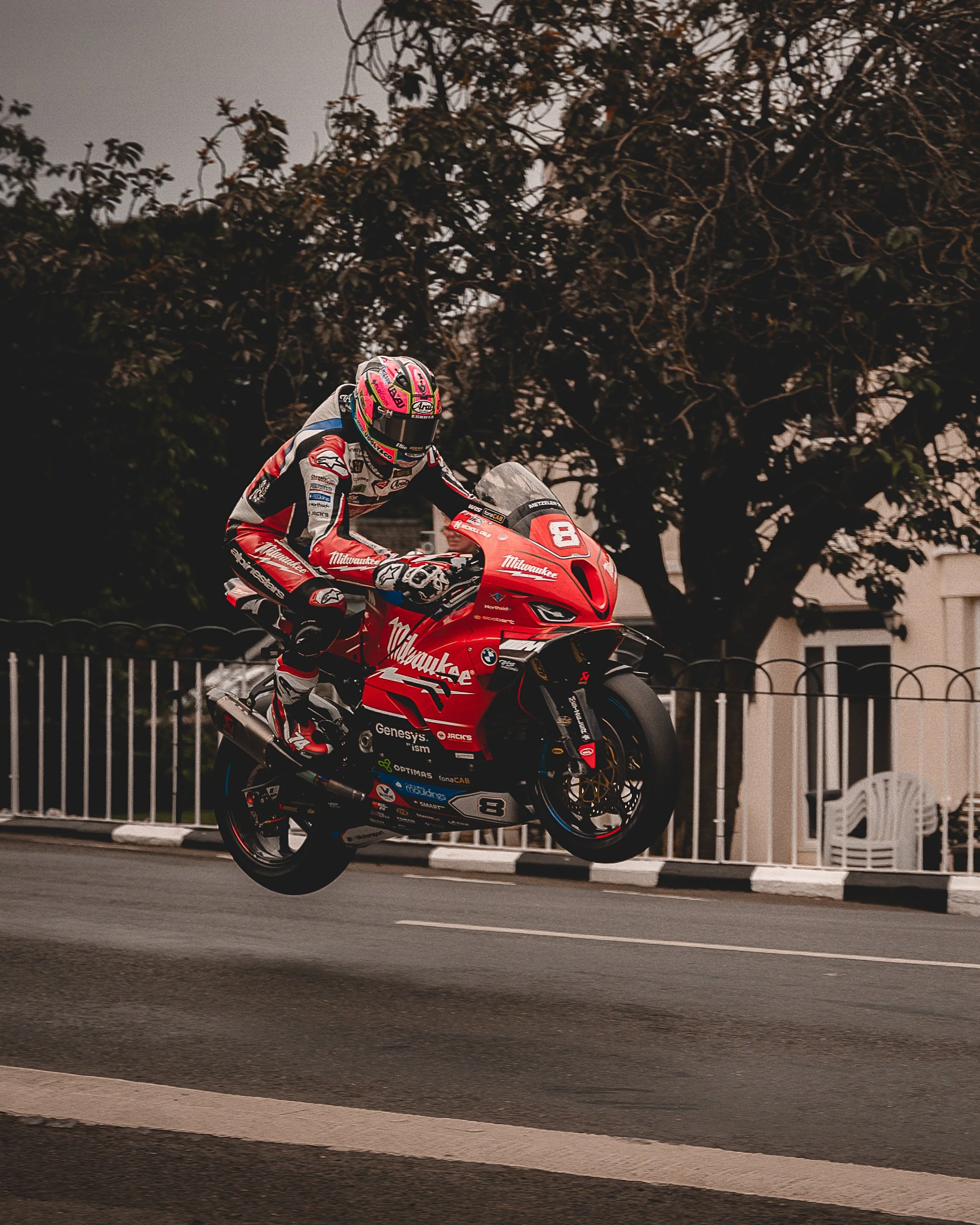 A motorcycle racer wearing a racing suit and helmet jumps over the road on a red motorcycle with racing decals, with trees and houses in the background.