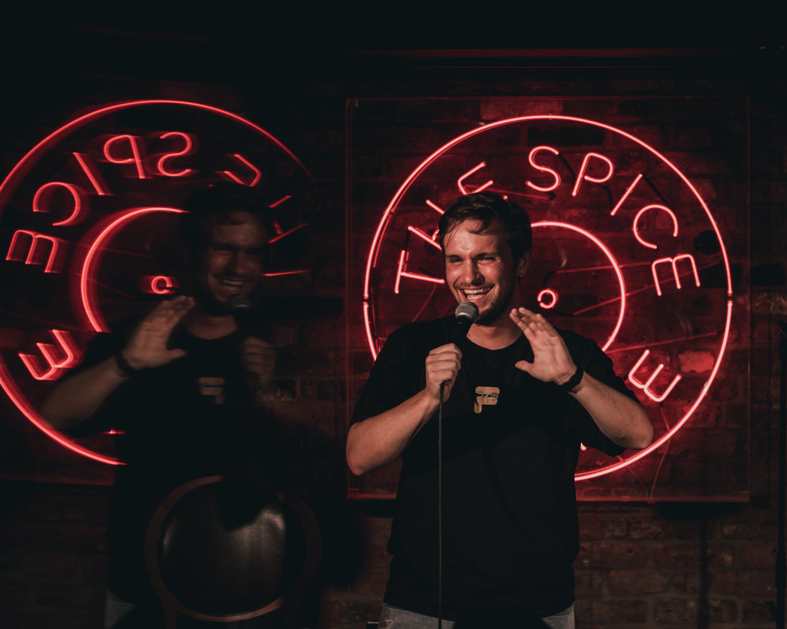 A man holds a microphone and laughs while speaking or performing in front of a red neon sign that reads 'THE SPIC'. His reflection is visible in the neon sign behind him, and the background appears to be a brick wall.