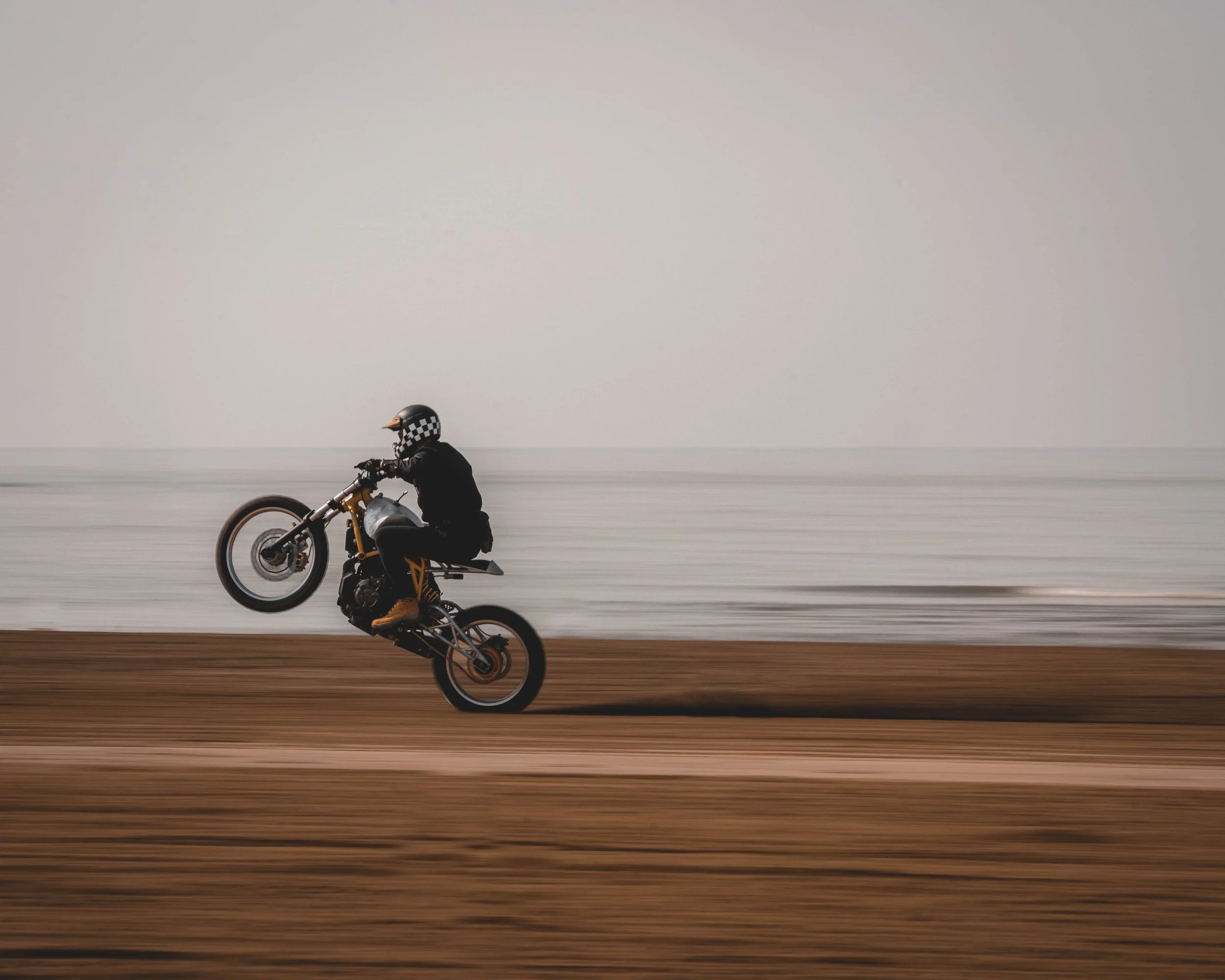 Person wearing a helmet performing a wheelie on a motorcycle on a beach.