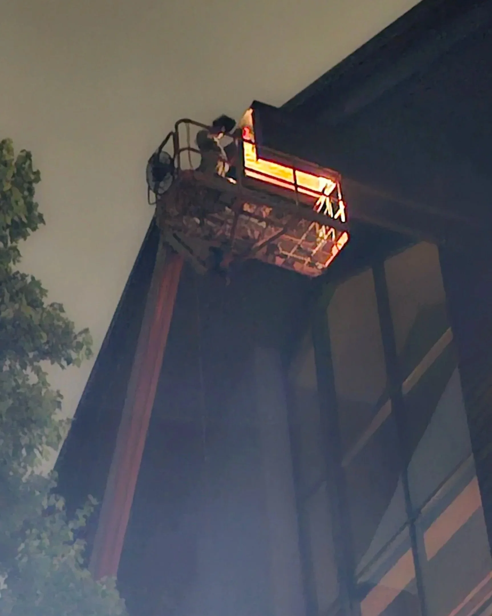 Nighttime scene showing a building with an emergency fire escape. The fire escape is illuminated with red and orange lights, appearing as glowing lines and shapes against the darker building facade. There is also a partial view of a tree with green leaves on the left side of the image.