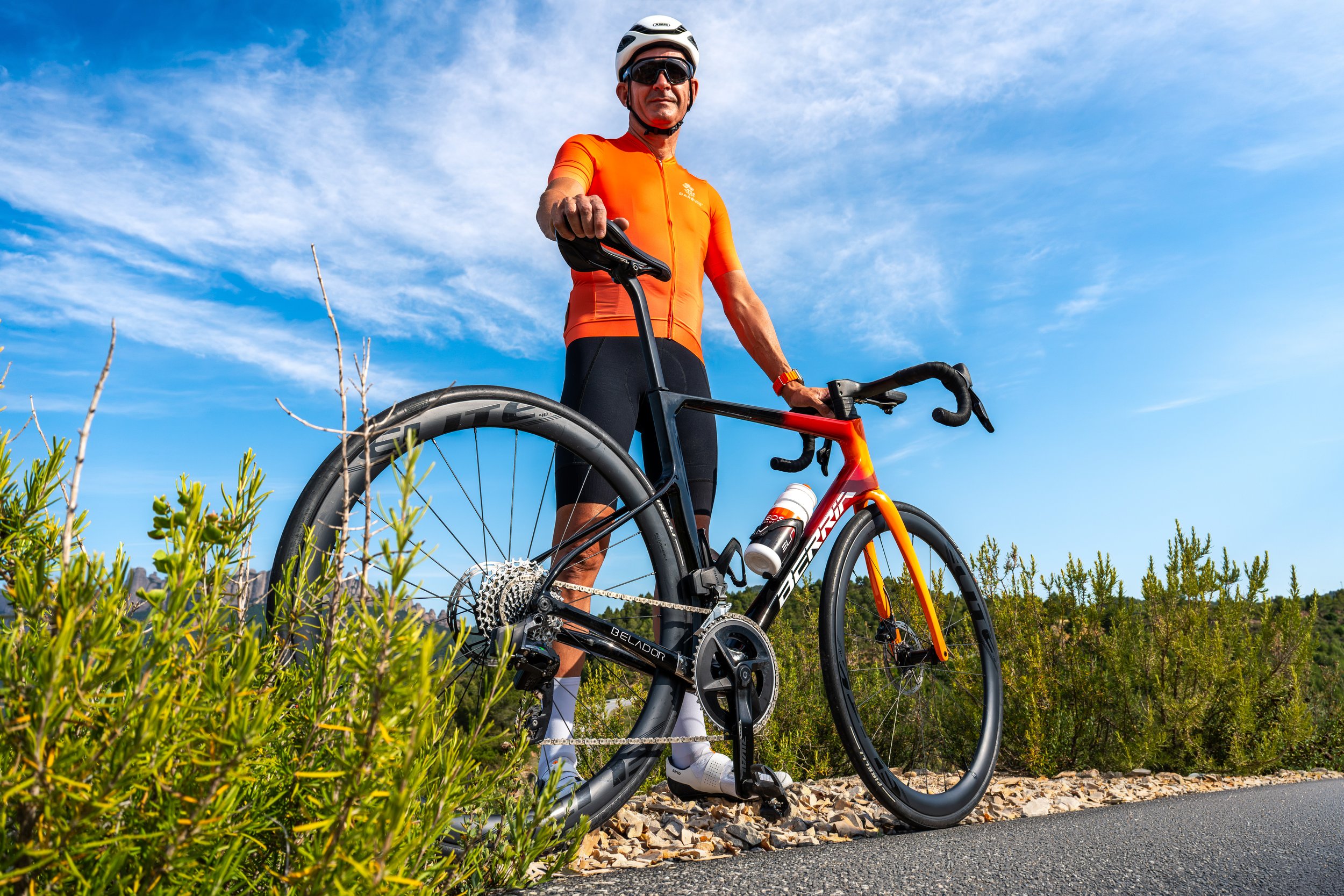 Hombre en equipo de ciclismo, con casco y gafas, posando con su bicicleta en un entorno natural con arbustos y cielo azul.
