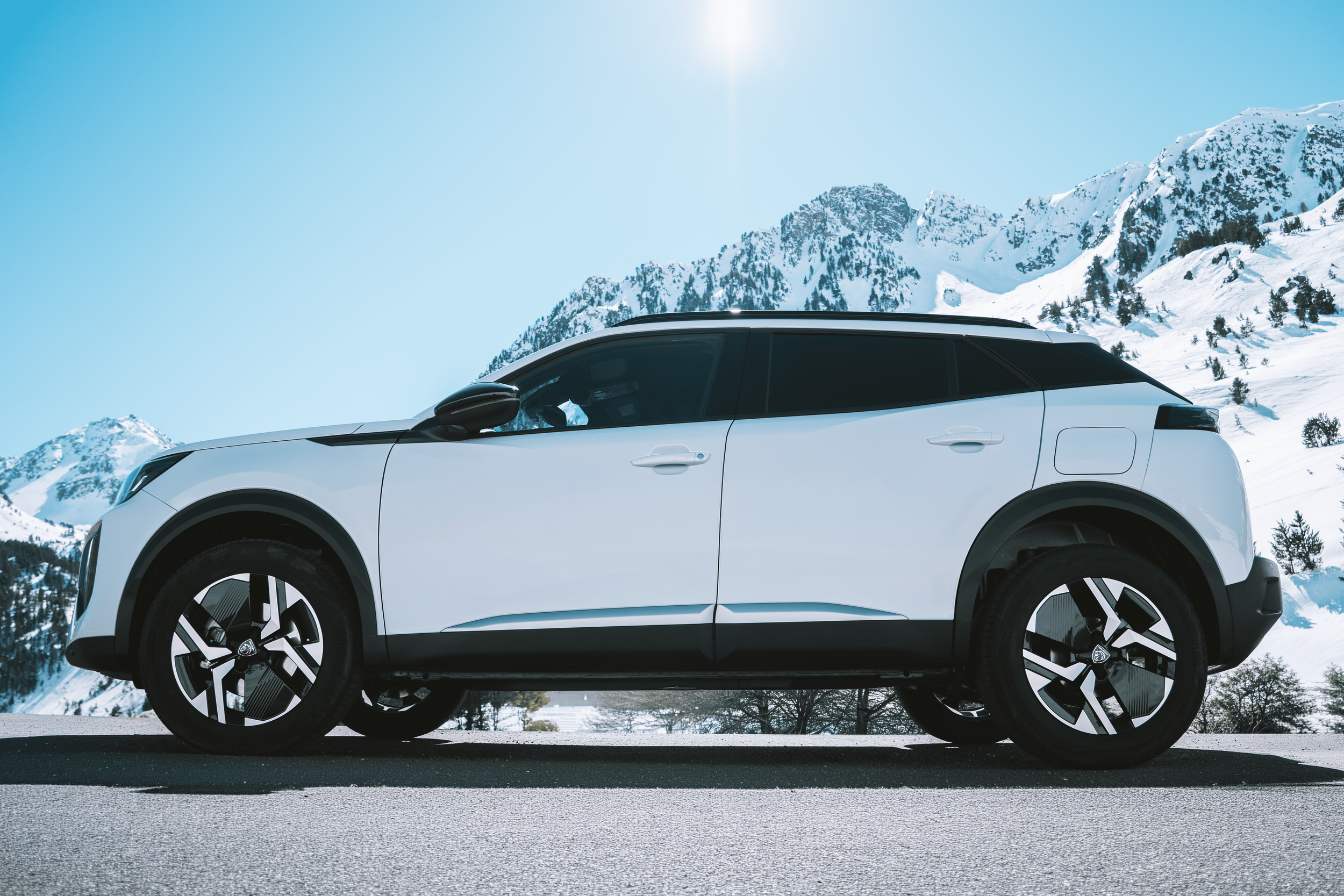 Auto deportivo de color blanco estacionado en un paisaje de montañas con nieve bajo un cielo despejado y sol brillante.