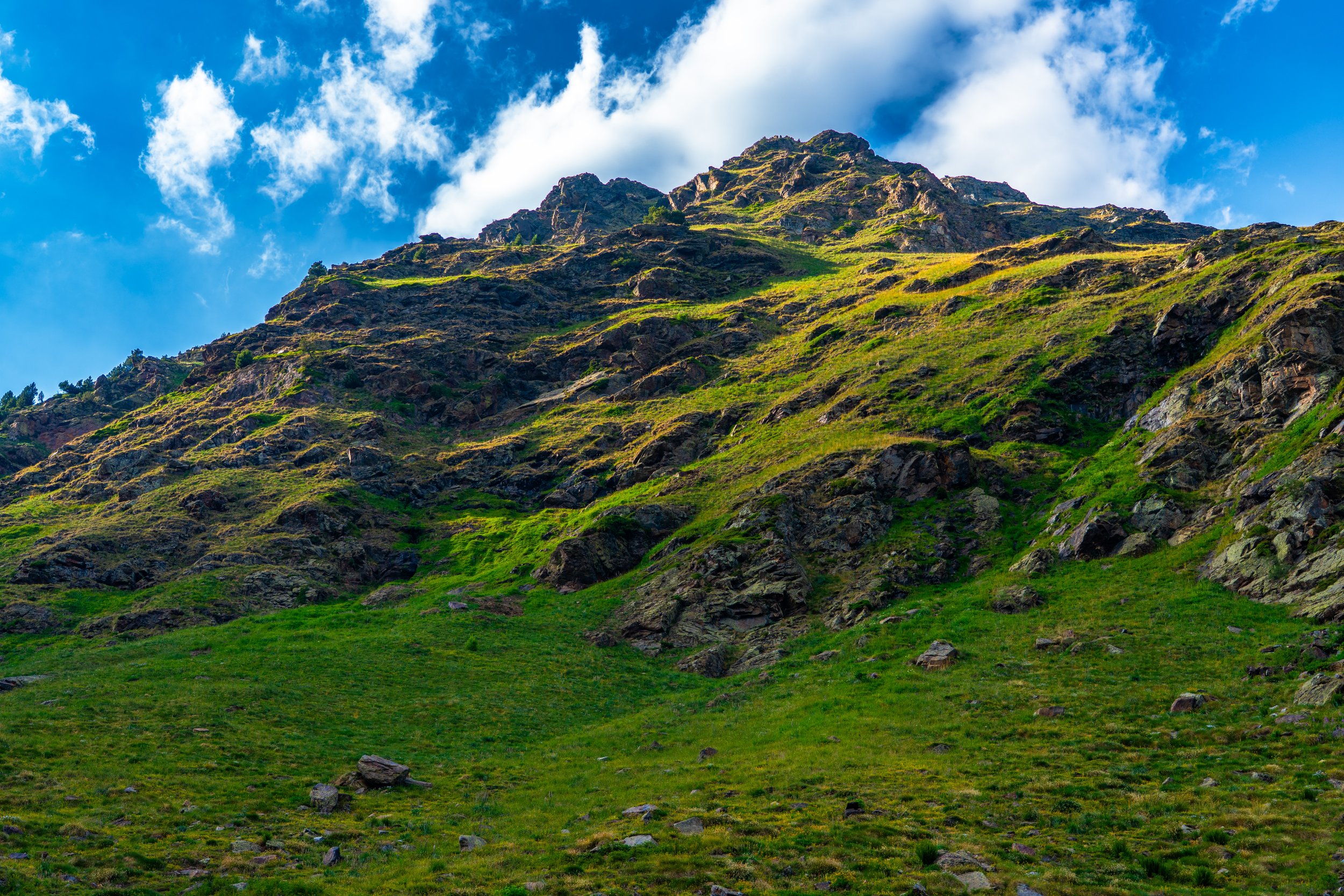 Montaña cubierta de hierba y rocas, con un cielo azul con nubes blancas.
