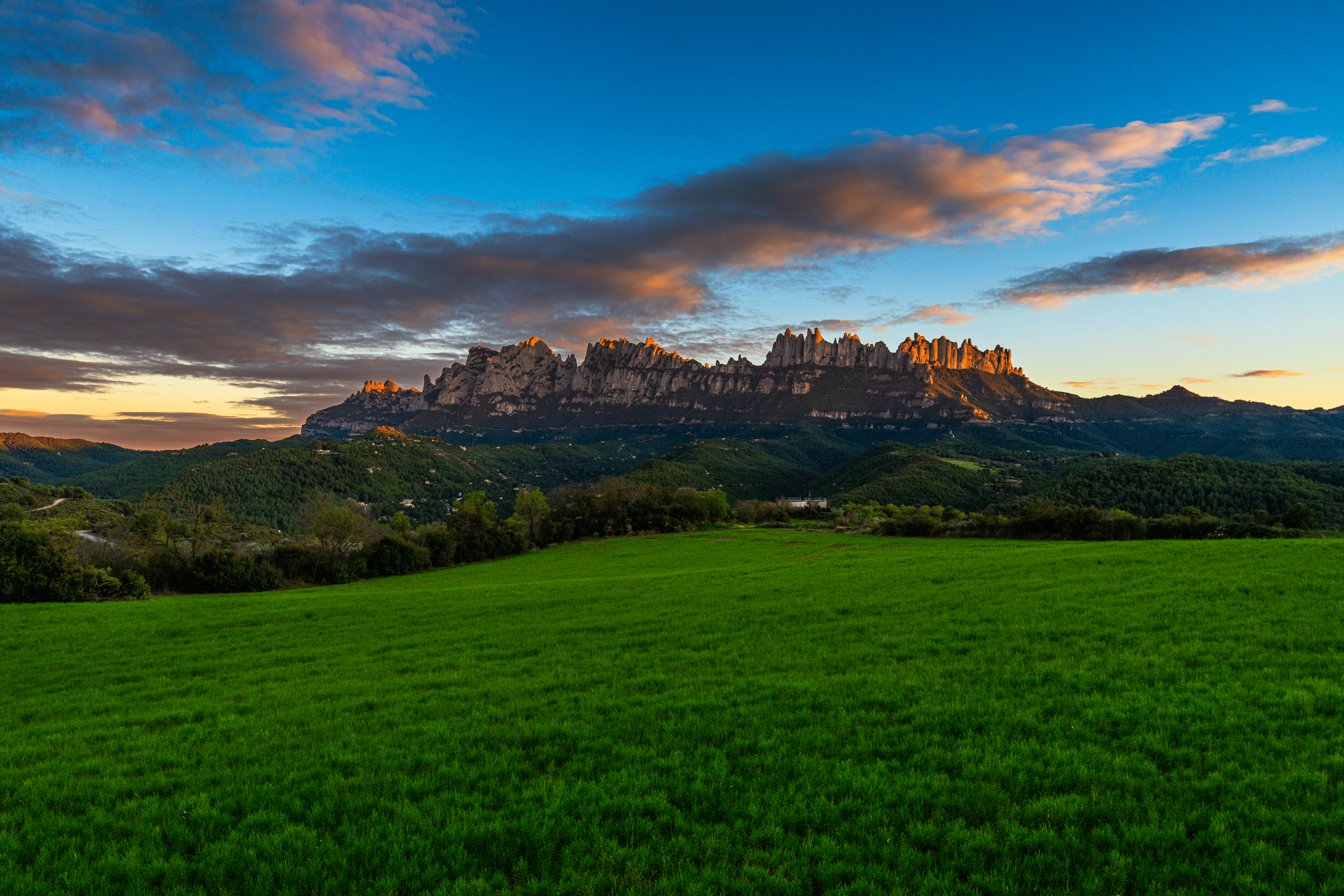 Montañas rocosas con picos escarpados iluminados por el atardecer, rodeadas de áreas verdes y cielo parcialmente nublado con nubes de color gris y rosa.