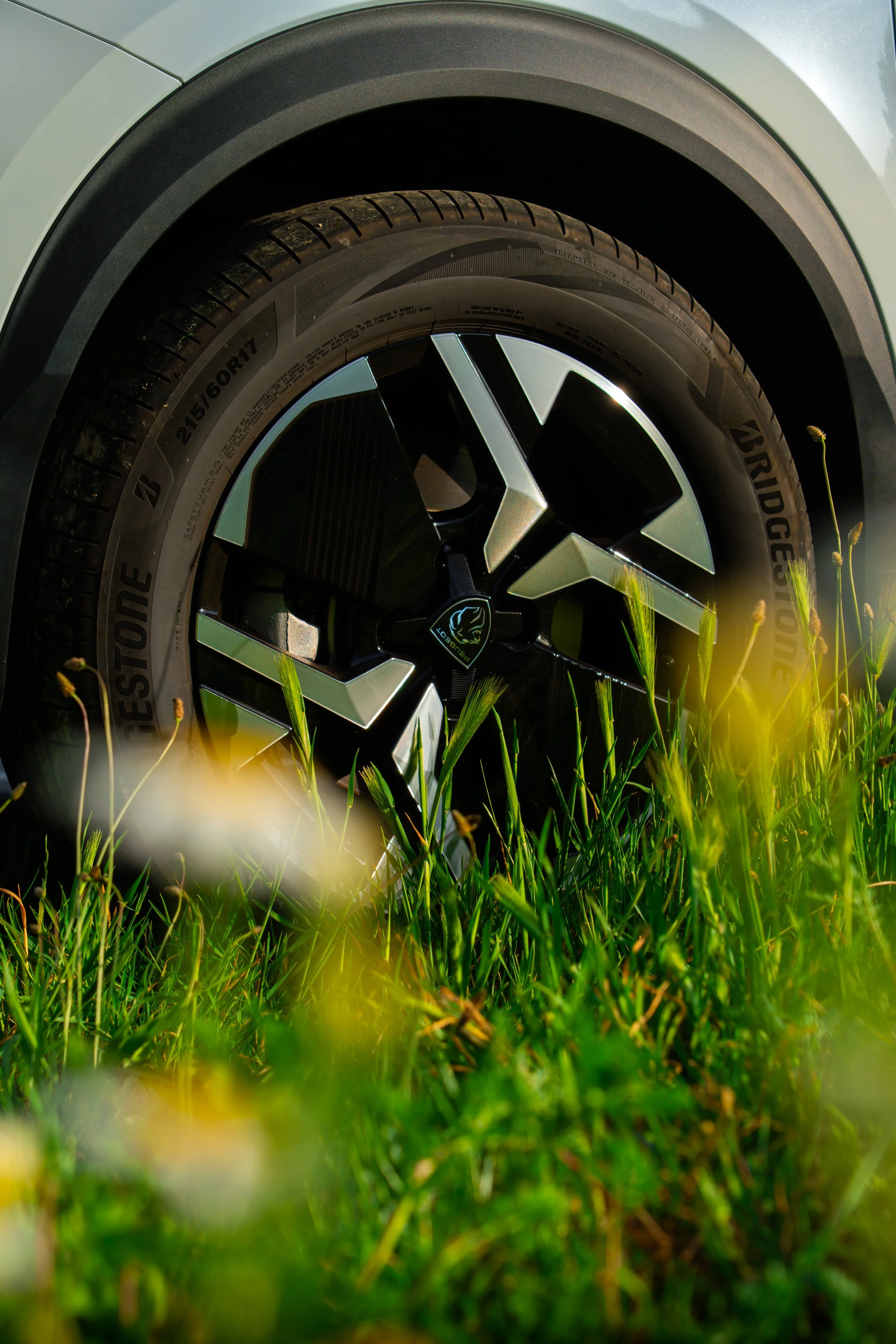 Cerca de la rueda de un automóvil que está estacionado en un campo de hierba verde con flores amarillas. La rueda tiene un neumático Bridgestone y una llanta de diseño moderno, con una calcomanía de un animal en el centro.