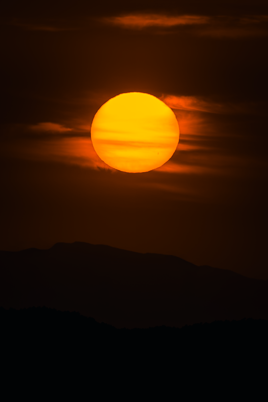Sol naranja brillante en un cielo oscuro, con nubes y montañas en el horizonte.