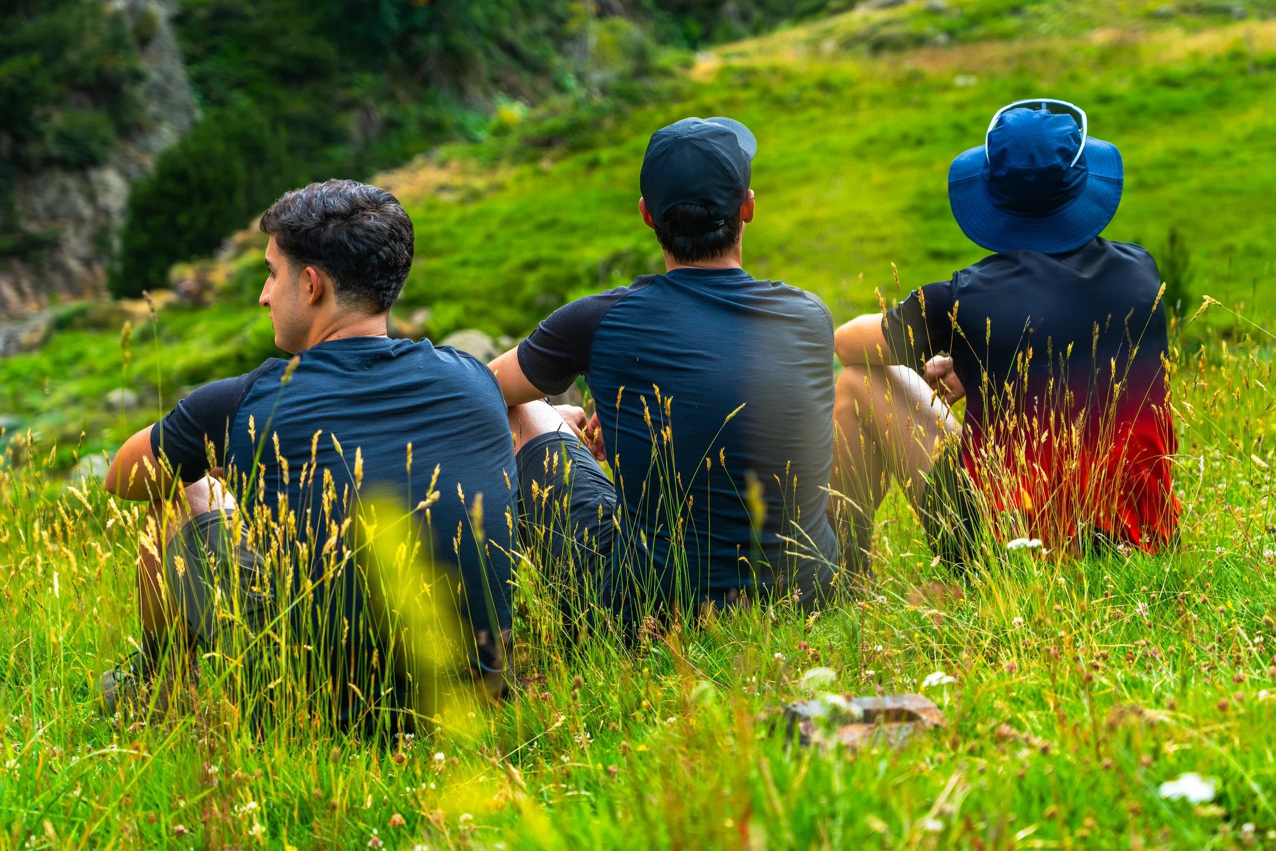 Tres personas sentadas en la hierba en un entorno de montaña verde, mirando hacia adelante.