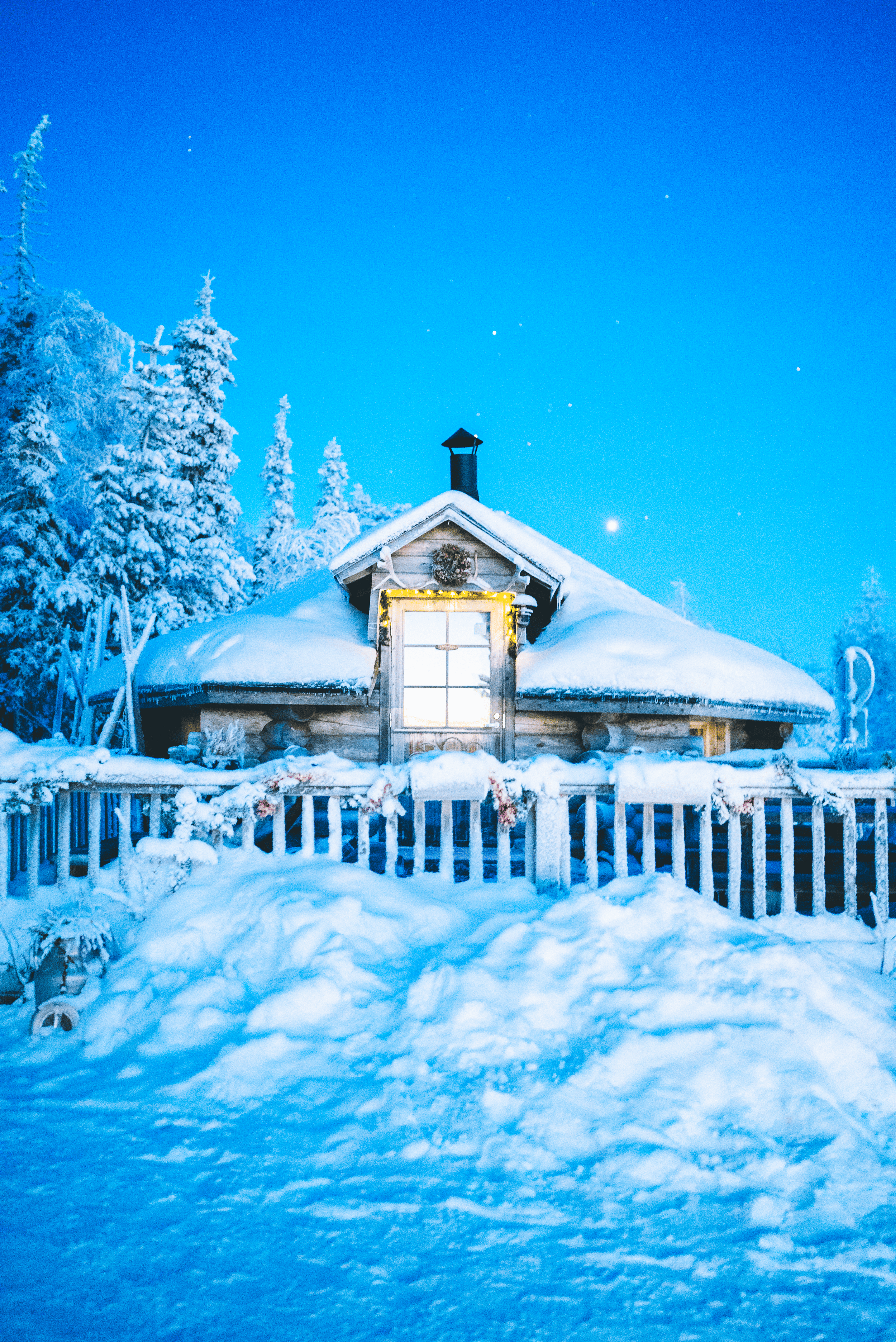 Una cabaña de madera cubierta de nieve, con ventanas iluminadas, rodeada de árboles cubiertos de nieve en un paisaje invernal nocturno con un cielo azul y estrellas.