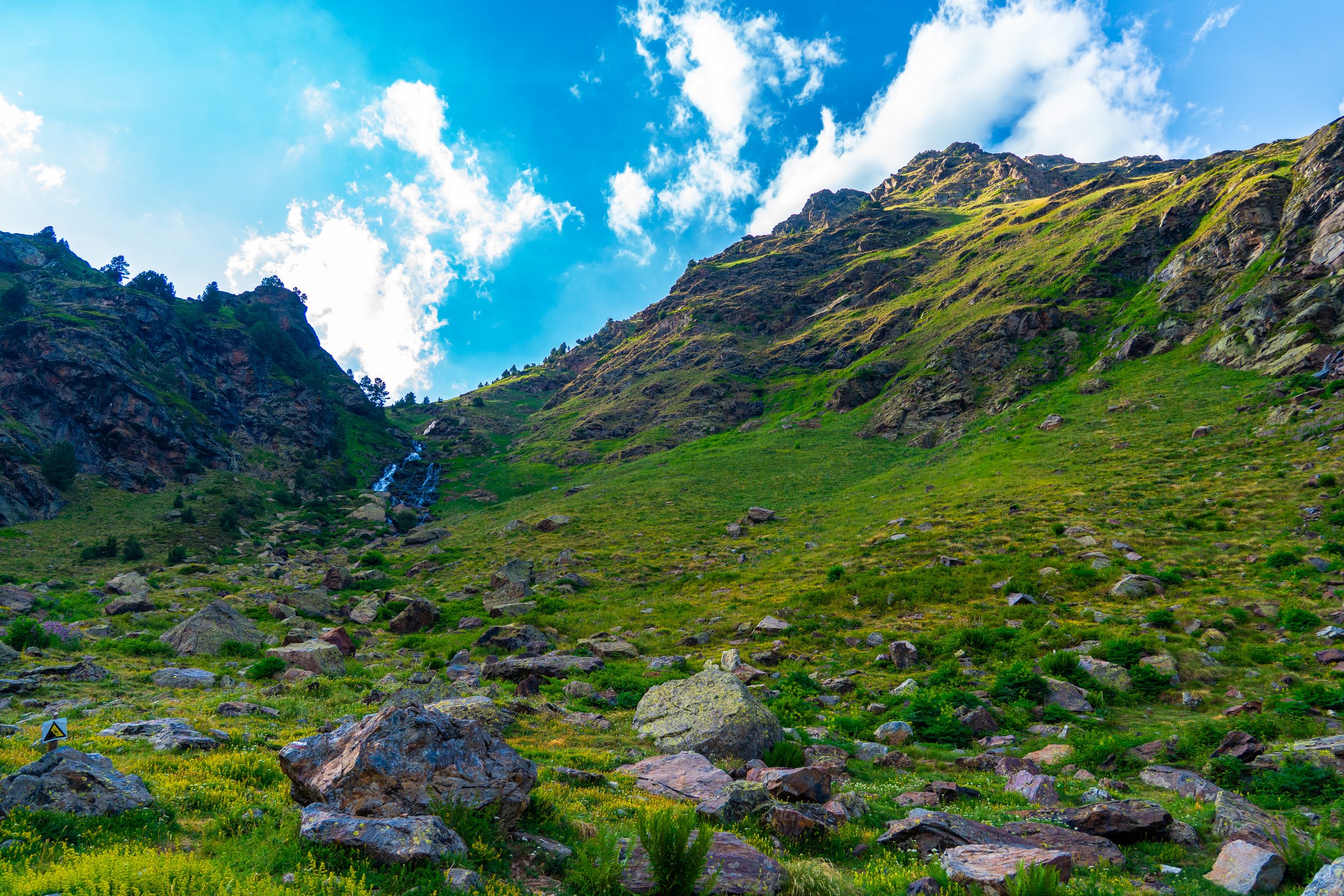Montaña verde con rocas grandes y un pequeño arroyo, cielo azul con nubes blancas.