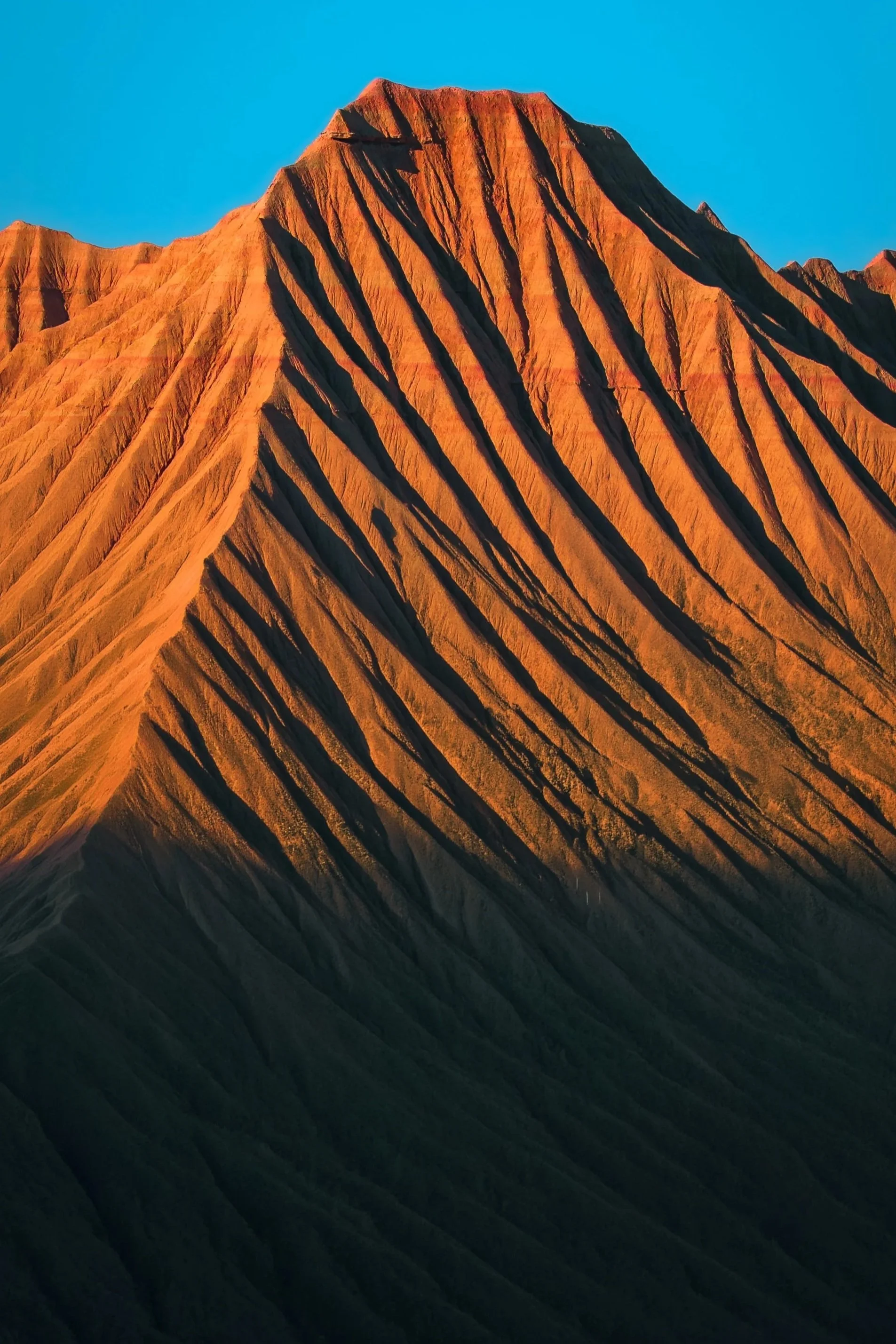 Montaña de color rojizo con formaciones escarpadas y líneas oscuras, bajo un cielo azul claro.