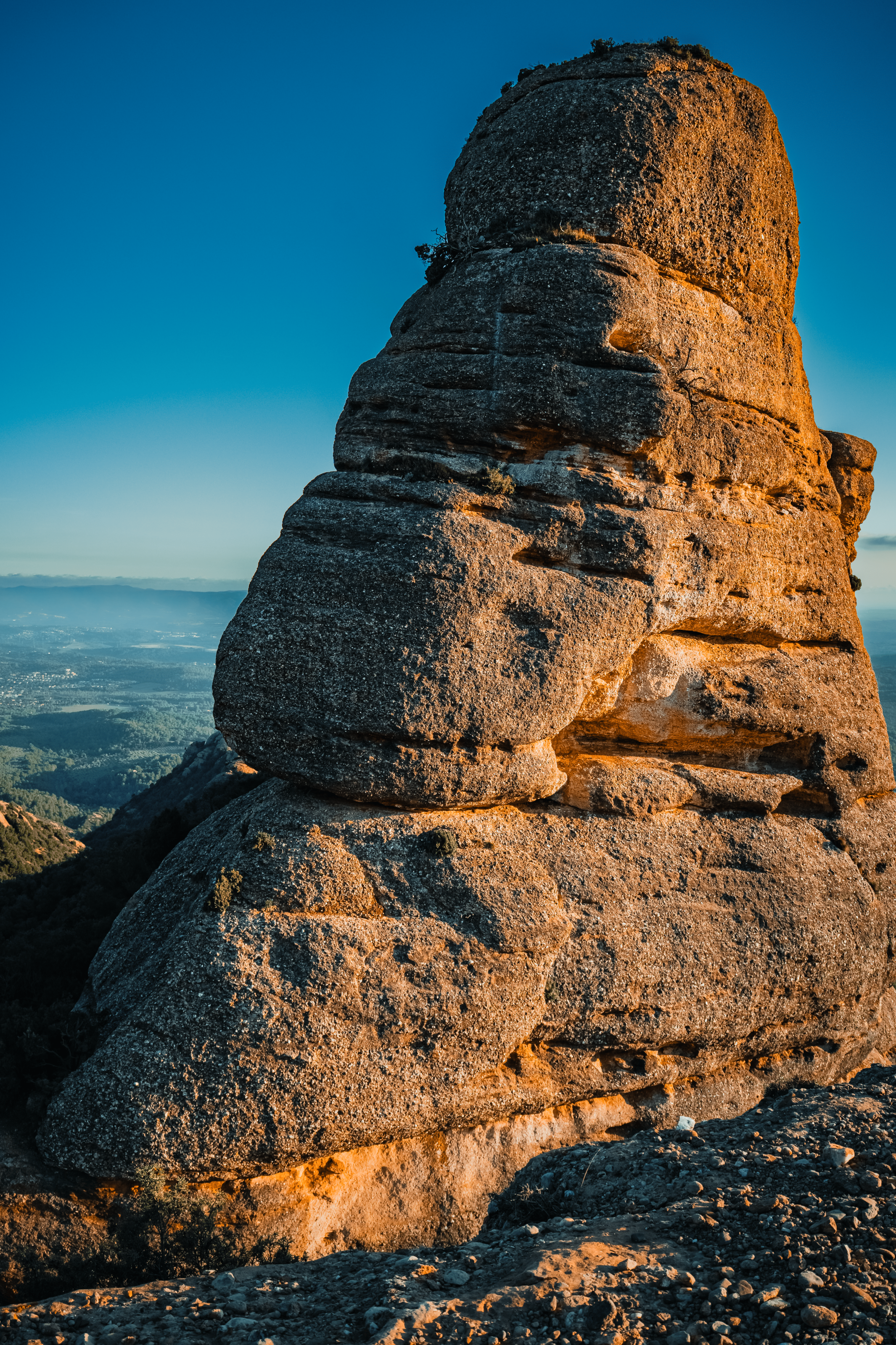 Caprichosa formación rocosa en una montaña con vista a un valle y cielo despejado al atardecer.