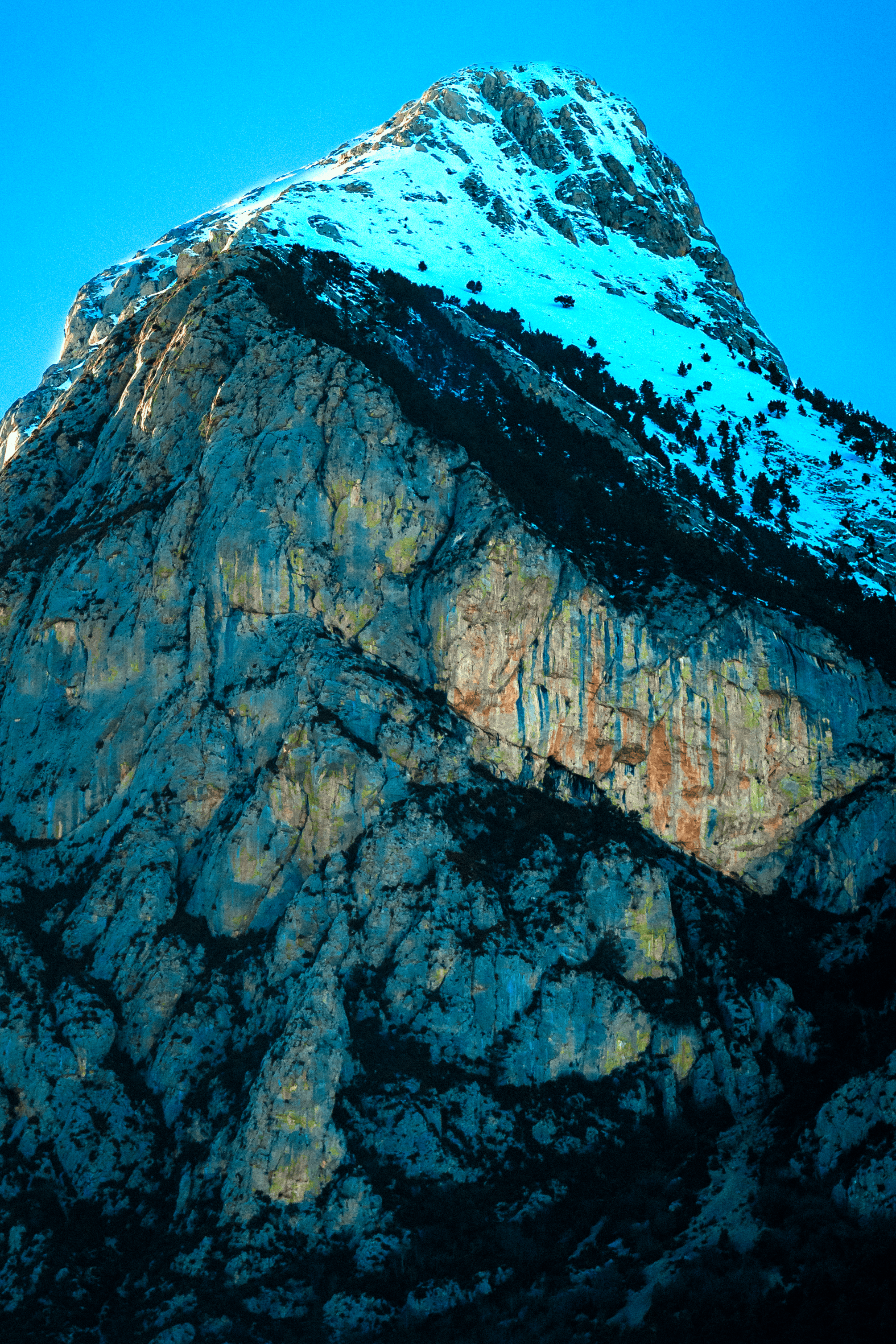 Una montaña nevada con rocas rojas y verdes en la base, bajo un cielo despejado y azul.