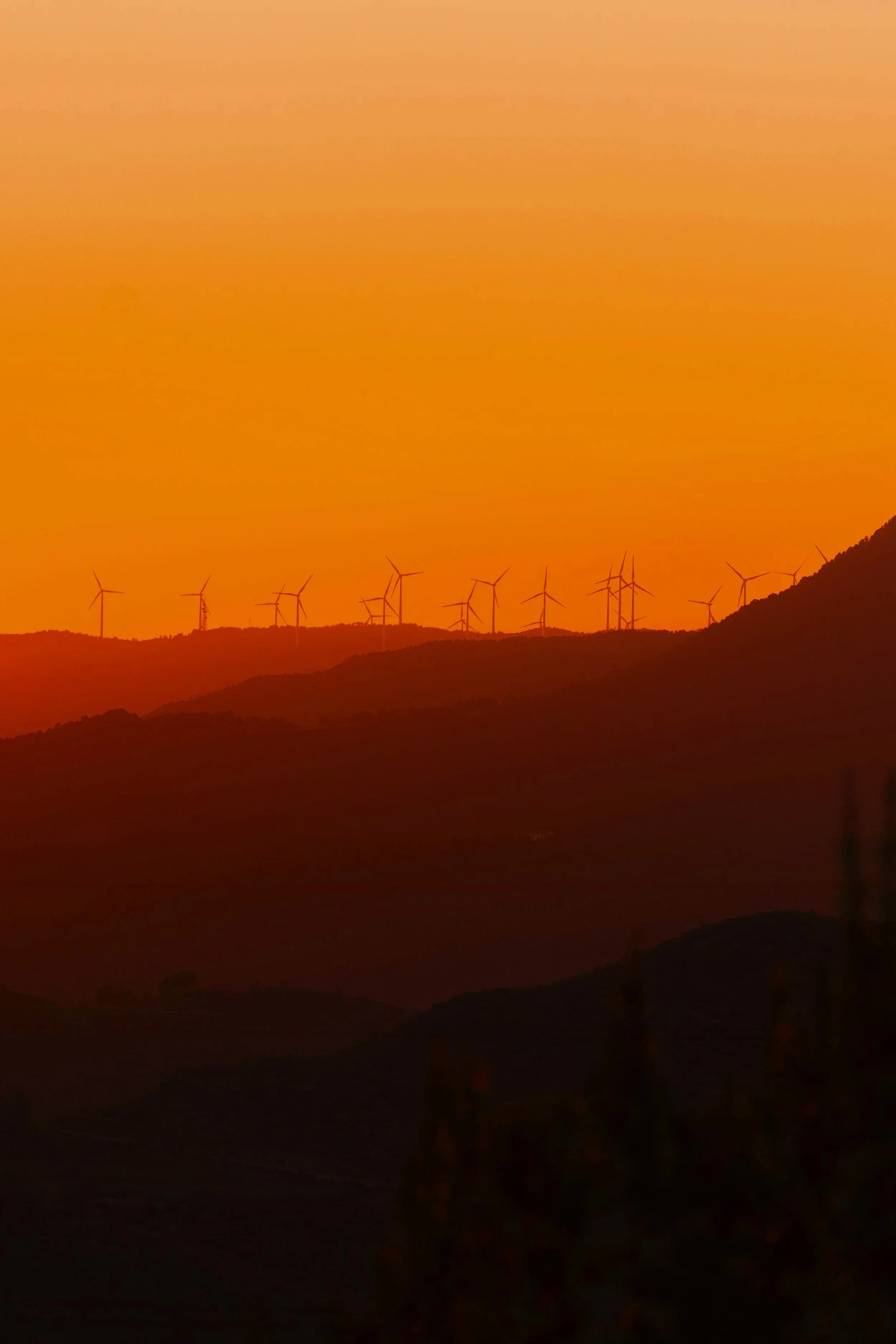 Paisaje montañoso con turbinas eólicas bajo un cielo anaranjado durante el atardecer.