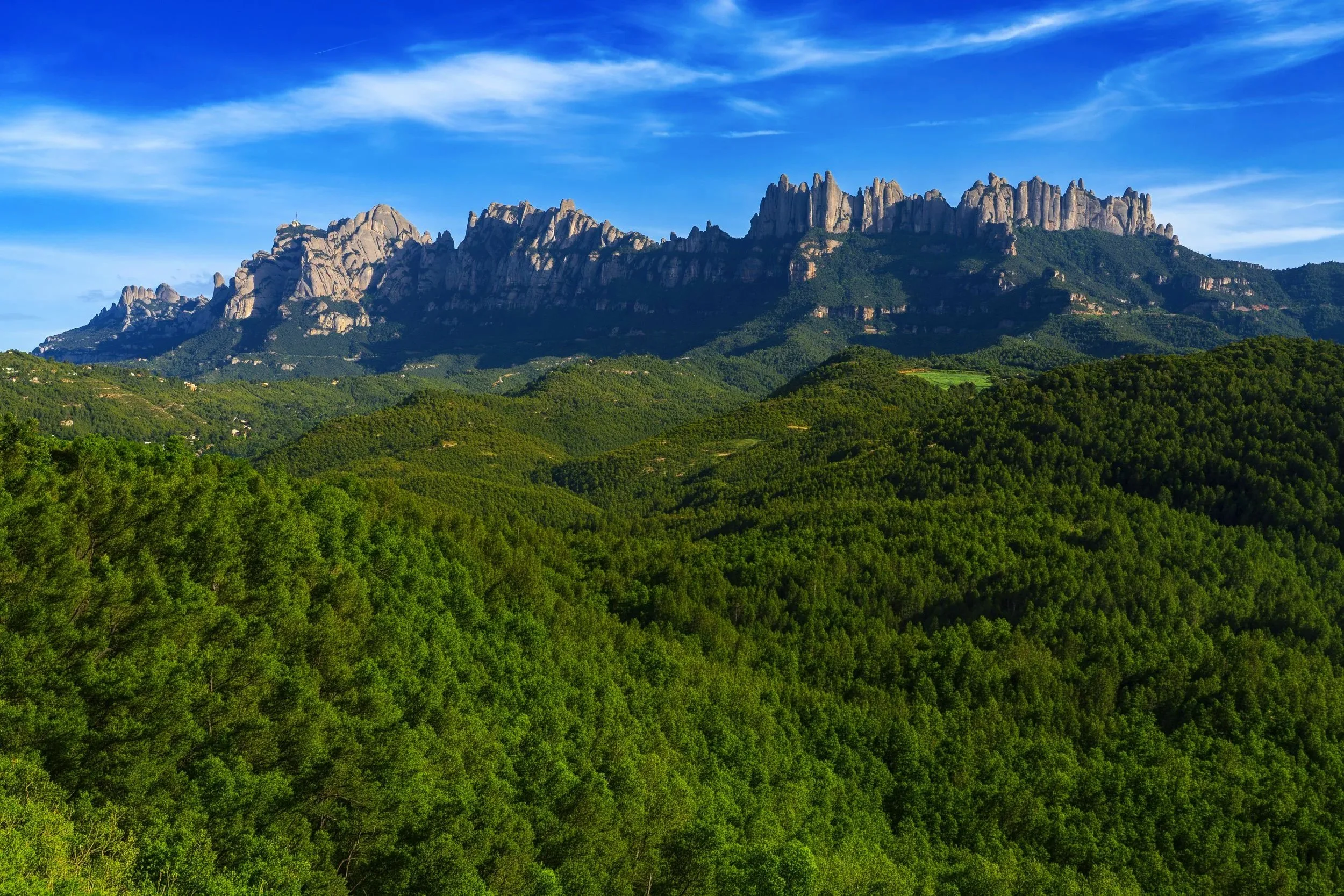Montañas rocosas con un cielo azul y nubes delgadas sobre un paisaje verde y frondoso.