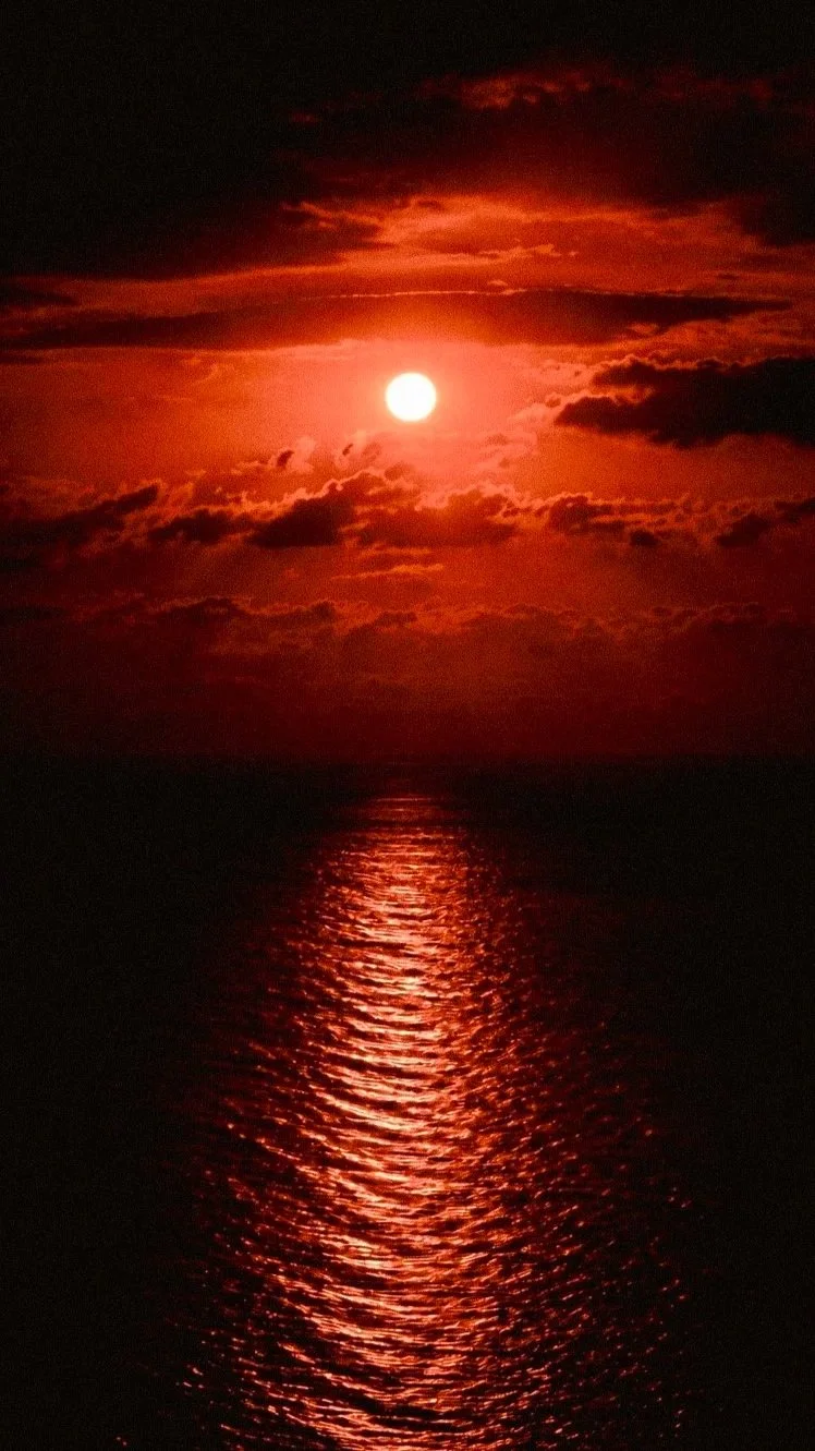 Atardecer con el sol brillante reflejándose en el agua del mar, con nubes oscuras y cielo en tonos rojos y naranjas.