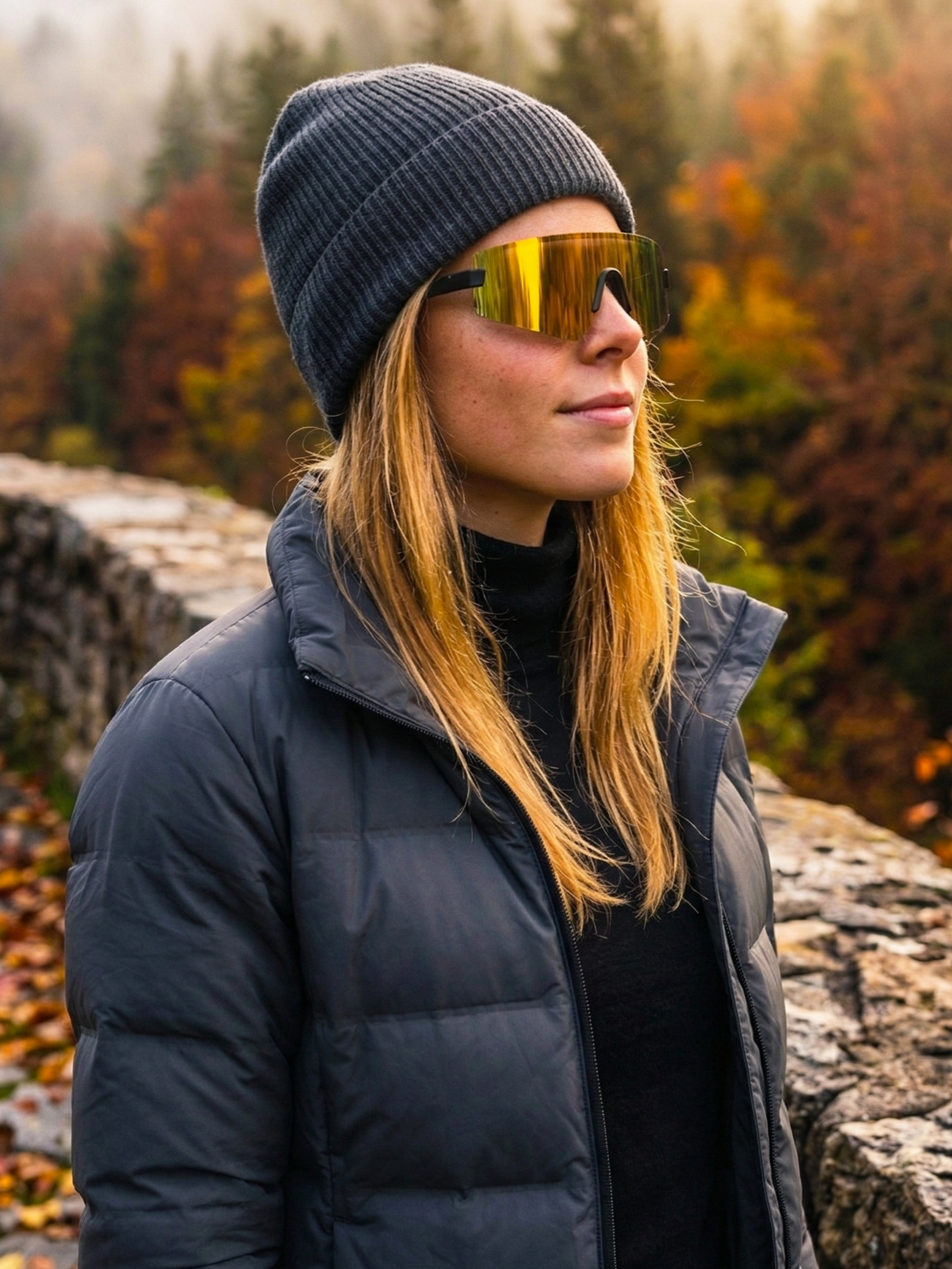 Mujer con gafas de sol, gorro gris y chaqueta, de perfil, en un entorno de bosque con hojas otoñales.
