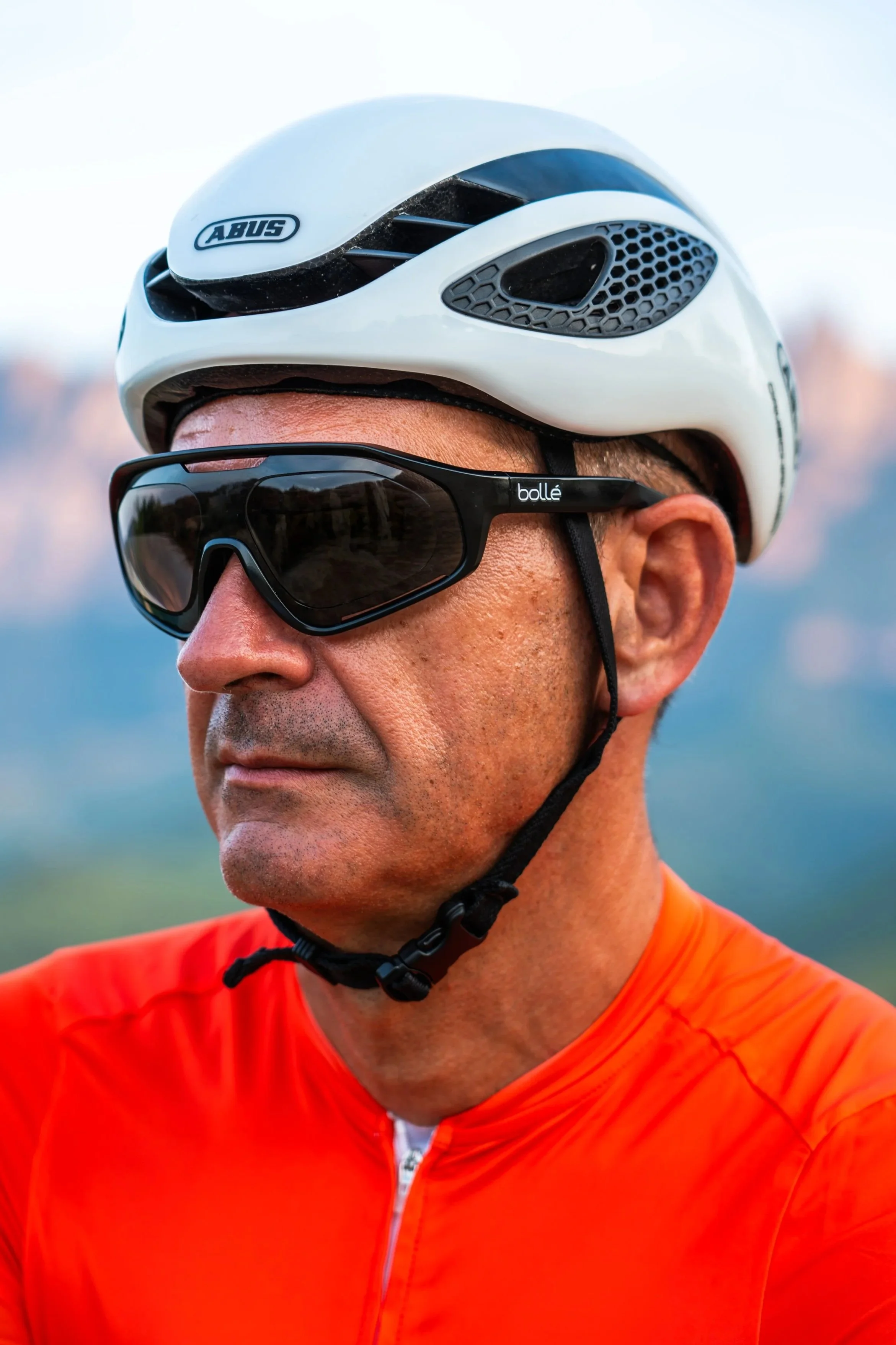 Hombre con gafas deportivas y casco de ciclismo de color blanco, vistiendo una camiseta naranja, en un entorno exterior