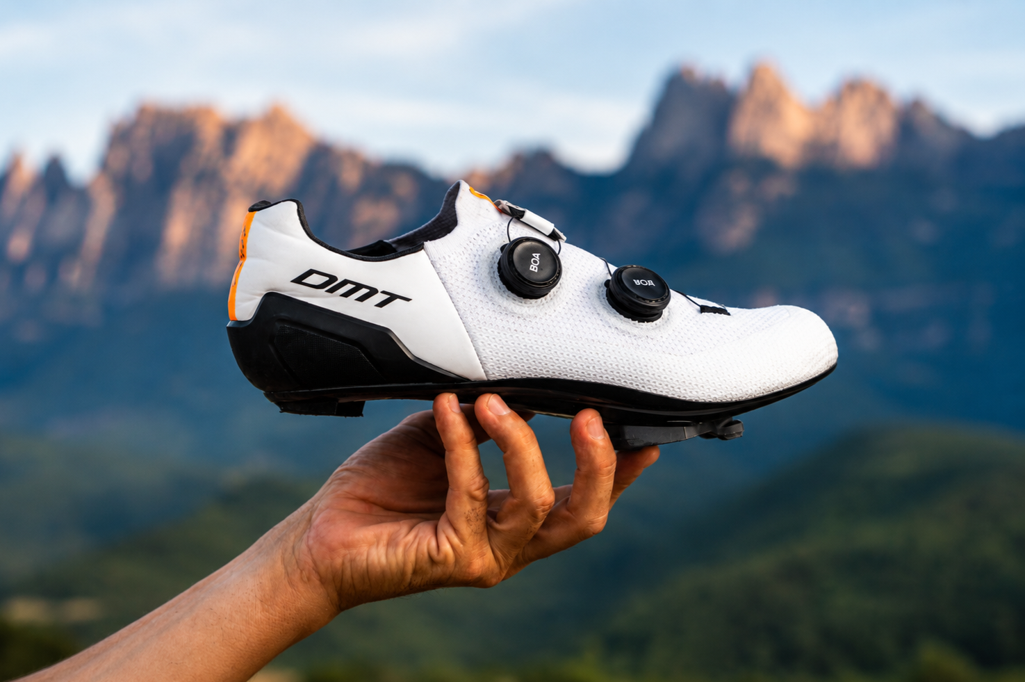 Zapato de ciclismo de carreras con calas y ajuste ajustado, marca DMT, en fondo montañoso y cielo azul.