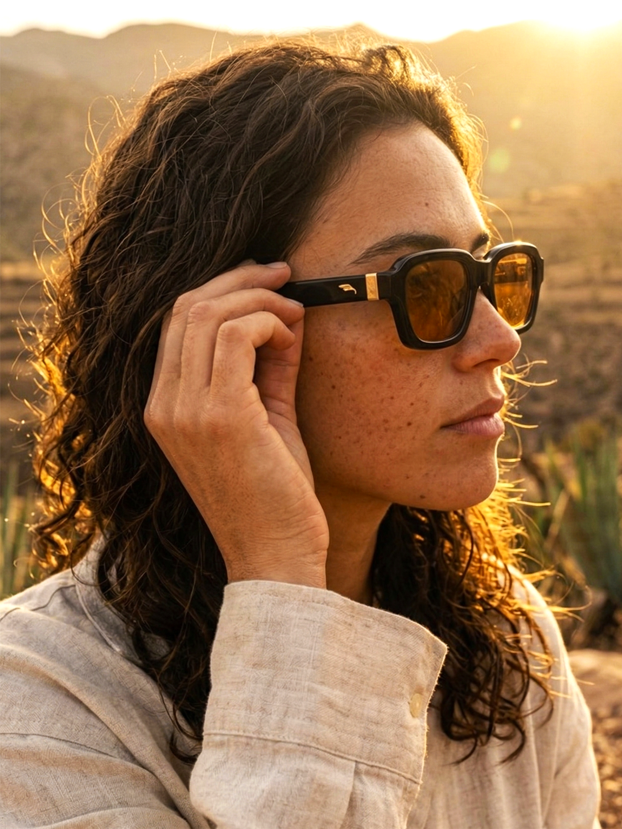 Mujer con cabello rizado y vestimenta clara ajustando sus gafas de sol en un campo al atardecer.