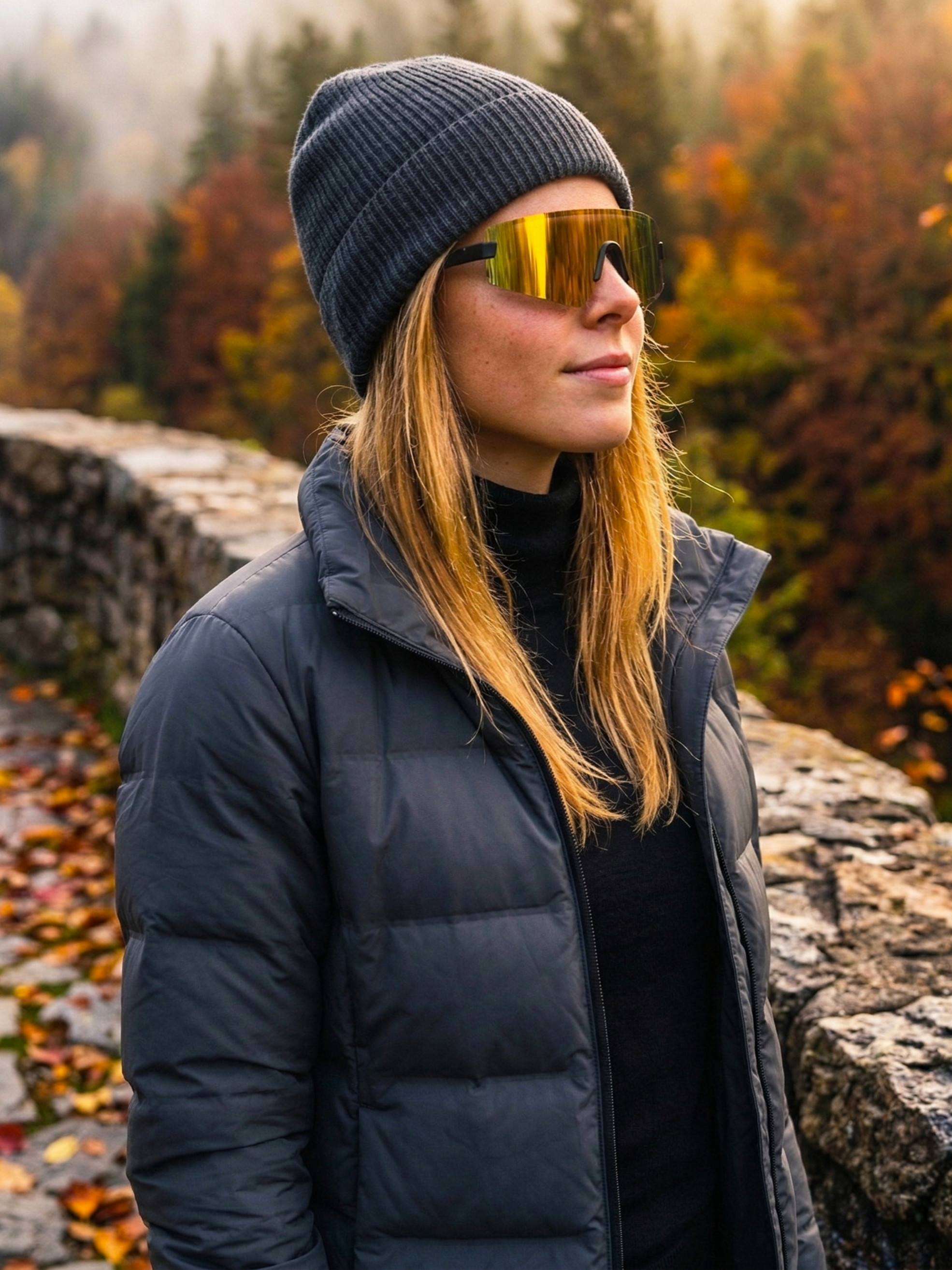 Una mujer con gafas de sol doradas, un gorro gris y una chaqueta negra de pie en un entorno natural con árboles otoñales y una cerca de piedra.