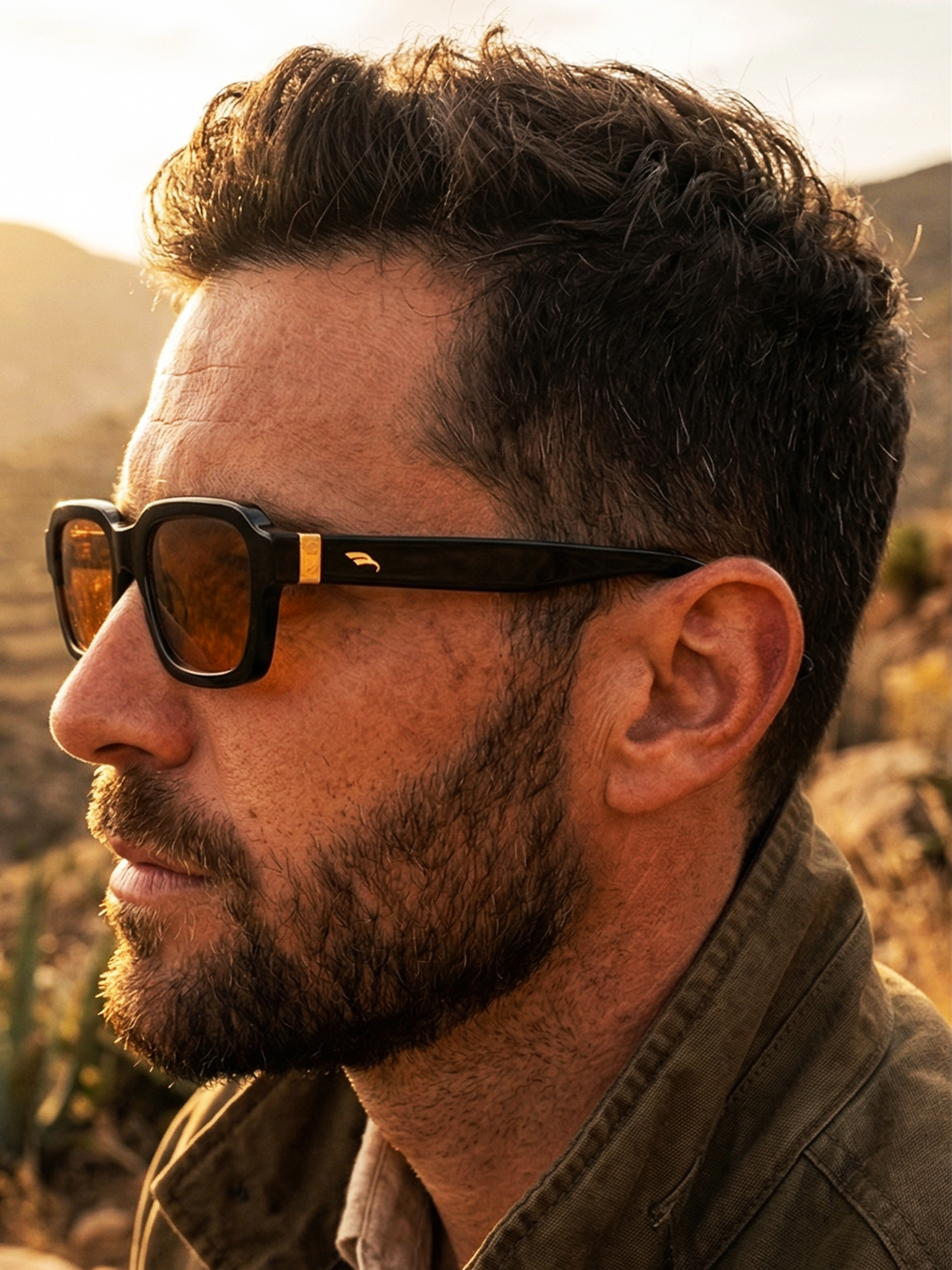 Retrato de un hombre con barba y barba, usando gafas de sol negras, en un entorno natural, al atardecer.